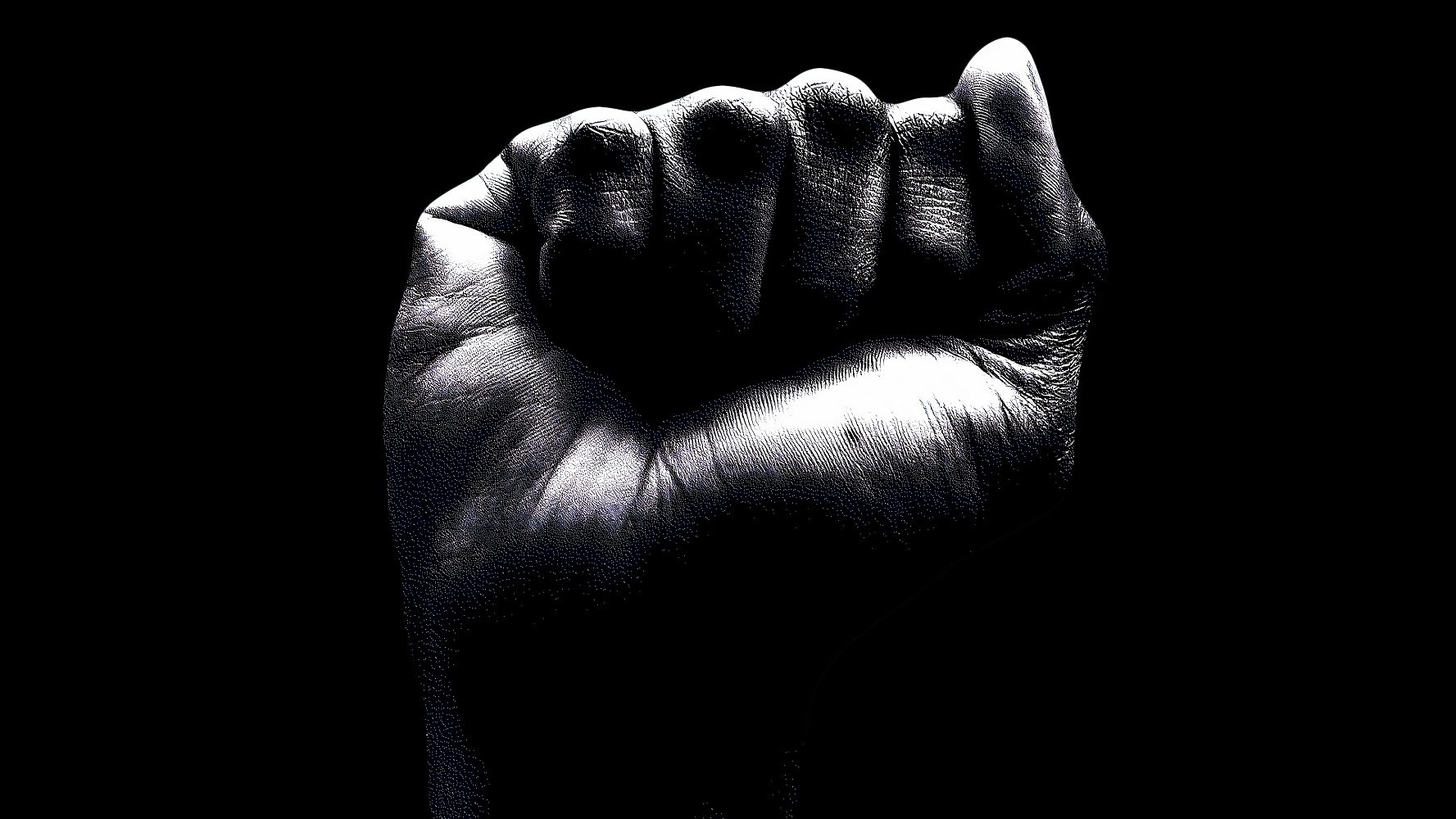L'image représente un poing fermé, vu de près sur un fond noir. La main est d'une teinte sombre, avec des détails visibles sur la peau, comme les lignes des empreintes digitales et les plis. Le poing, symbolisant souvent la force ou la résistance, est levé de manière à exprimer la détermination. Les contours de la main sont nets, mettant en avant la texture et la forme. L'arrière-plan noir accentue la présence du poing, créant un contraste fort et saisissant.