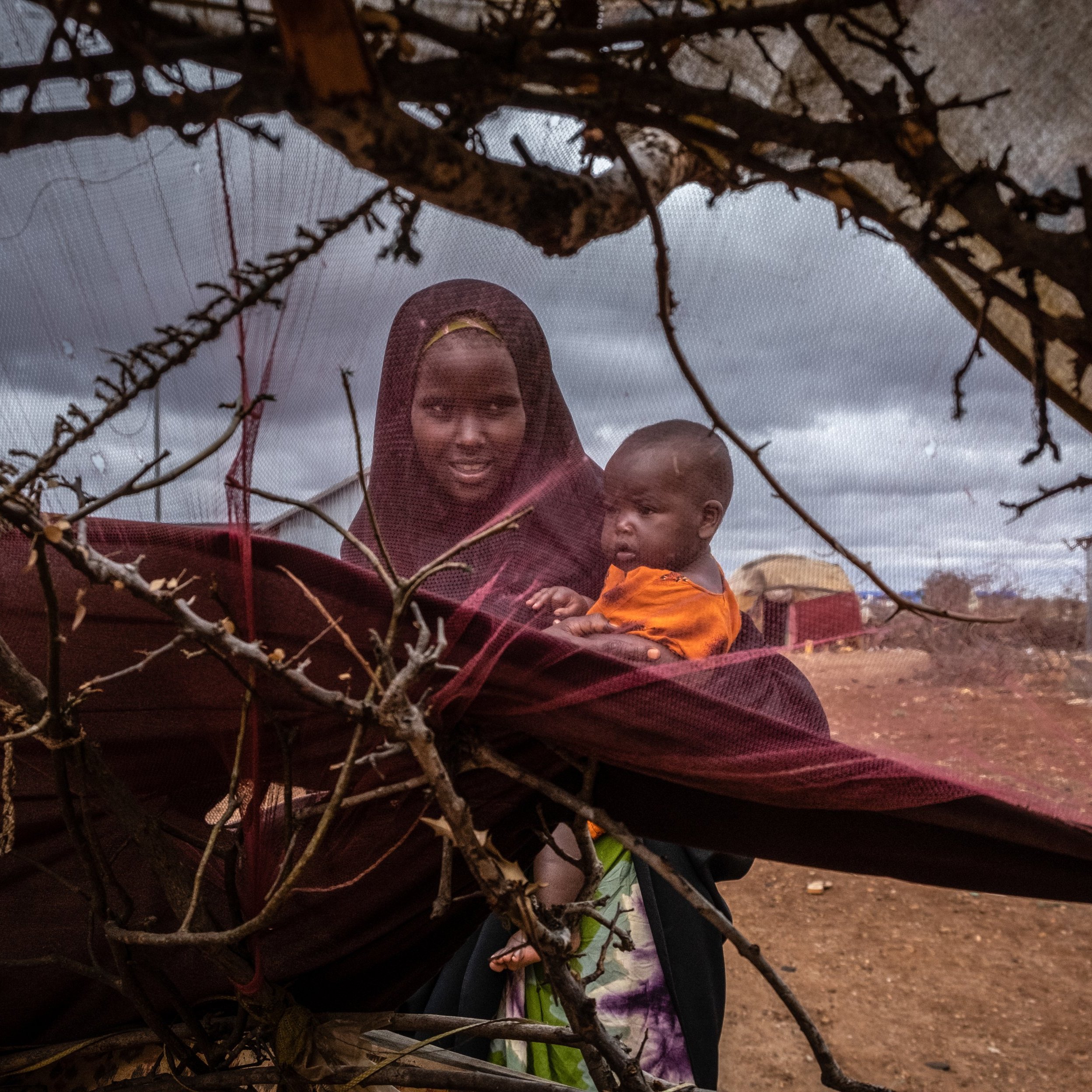 L'image montre une femme et un enfant au sein d'un environnement difficile. La femme, vêtue d'un voile sombre et d'habits traditionnels, tient l'enfant dans ses bras. L'enfant, portant un vêtement orange, semble curieux et regarde autour de lui. Ils sont entourés de branches et de tissus, ce qui forme une sorte d'abri rudimentaire. Le sol est poussiéreux et l'atmosphère est chargée d'une lumière diffuse, suggérant un ciel nuageux. Cette scène évoque la résilience et le lien fort entre la mère et son enfant dans un contexte de précarité.