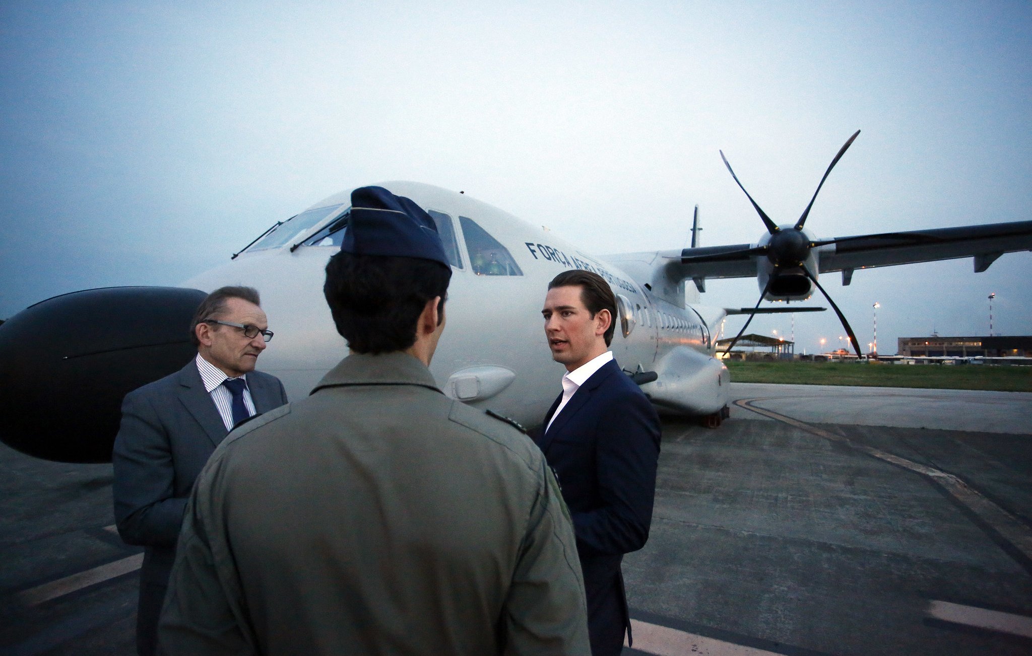 L'image représente un groupe de trois hommes se tenant près d'un avion sur une piste d'atterrissage. L'avion a des hélices et semble être un appareil de taille moyenne. L'éclairage est doux, indiquant qu'il peut être tôt le matin ou tard en soirée. Un homme en costume noir discute avec un autre homme habillé d'une tenue militaire. Le troisième homme, portant des lunettes, semble écouter la conversation. L'environnement est urbain et l'aéroport est visible en arrière-plan avec quelques lumières. L'atmosphère est professionnelle et engageante.