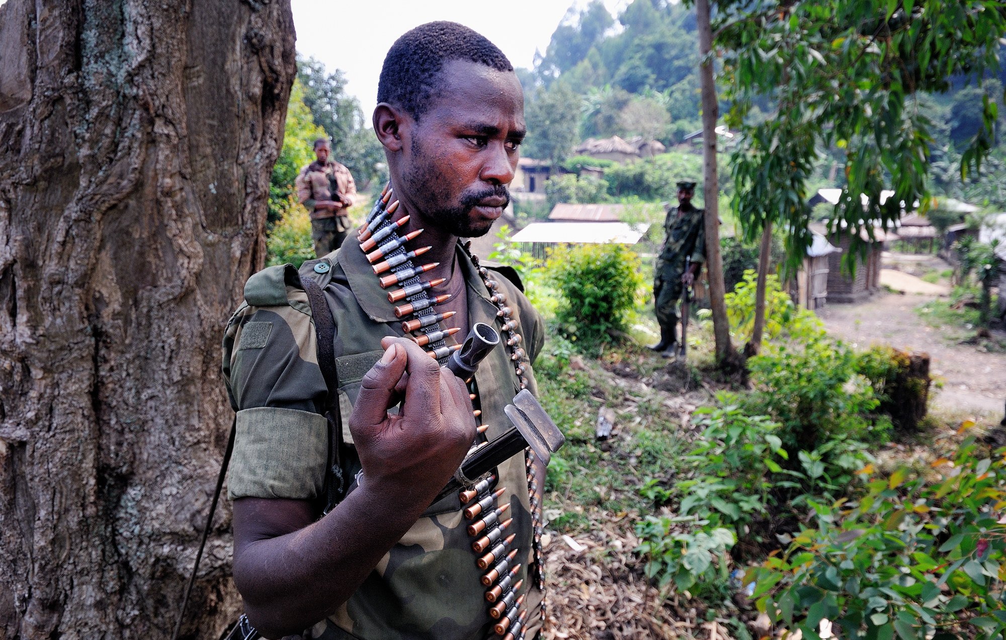 L'image montre un soldat debout près d'un arbre, portant un uniforme militaire camouflage. Il a une ceinture de munitions autour de son torse, et il tient une arme à la main. Son expression est sérieuse, indiquant la concentration et la vigilance. En arrière-plan, d'autres soldats sont visibles, et le paysage est verdoyant, avec des habitations simples au loin. L'atmosphère dégage une impression de tension en raison du contexte militaire.