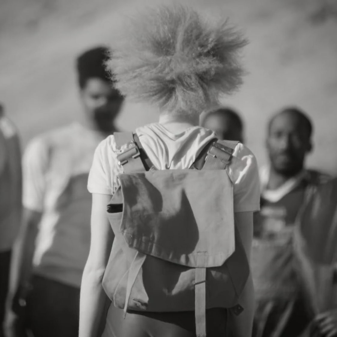 L'image présente un groupe de personnes, avec au premier plan une personne aux cheveux crépus et blonds, vue de dos. Elle porte un sac à dos. L'arrière-plan est flou, mais on devine des montagnes ou des collines. Les autres individus, qui se tiennent plus loin, ont des expressions faciales sérieuses et semblent réunis dans une conversation ou une interaction. L'atmosphère de l'image est en noir et blanc, ce qui accentue les contrastes entre les différentes textures et les attitudes des personnages.