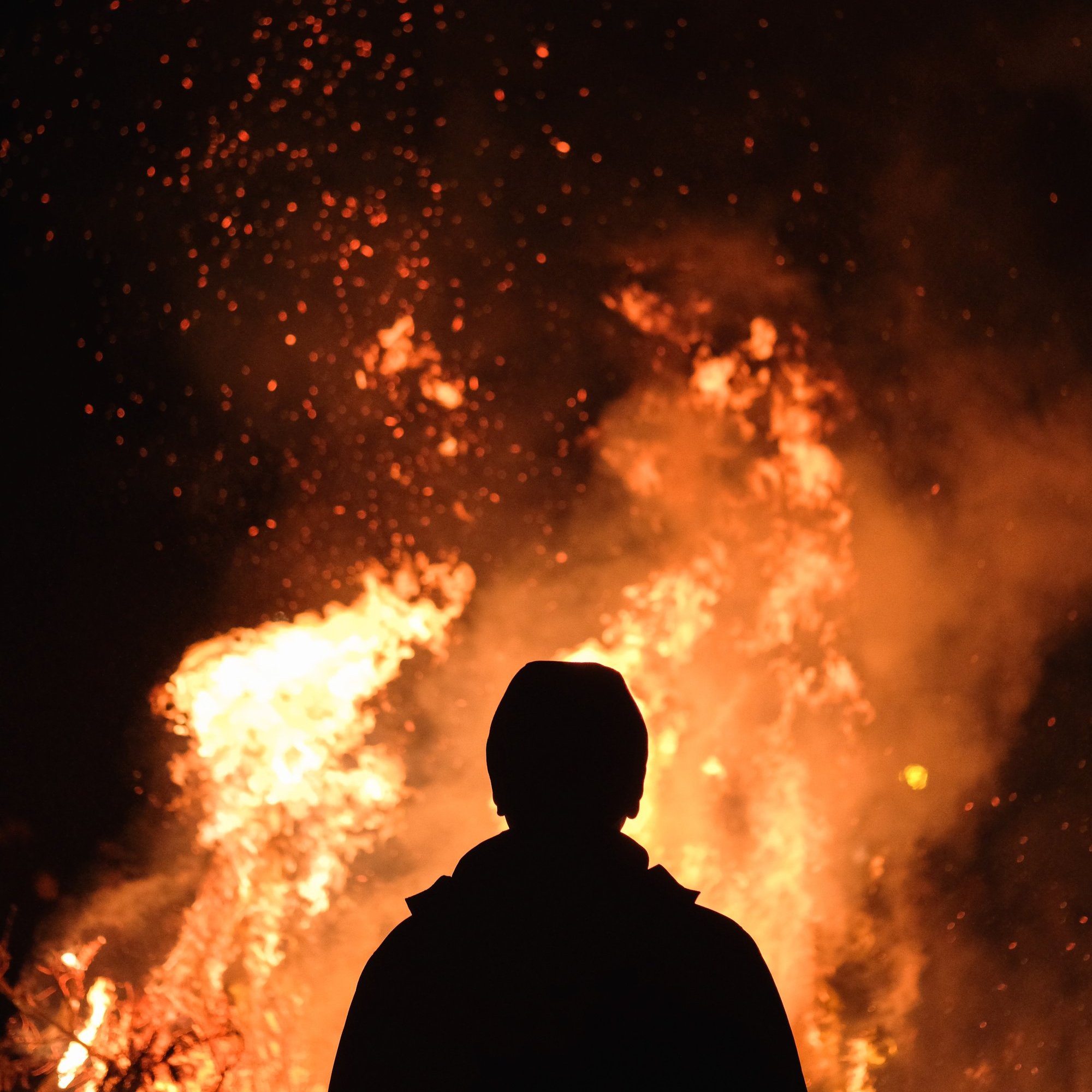 L'image montre une scène nocturne où une personne se tient de dos, face à un grand feu. Les flammes dansent avec éclat, projetant une lumière vive et chaleureuse dans l'obscurité environnante. Des étincelles s'élèvent en spirales, créant une ambiance à la fois magique et intense. La silhouette de la personne est sombre, contrastant avec les couleurs vives de l'incendie, ce qui permet de ressentir la chaleur émanant du feu et l'atmosphère captivante qui l'entoure.