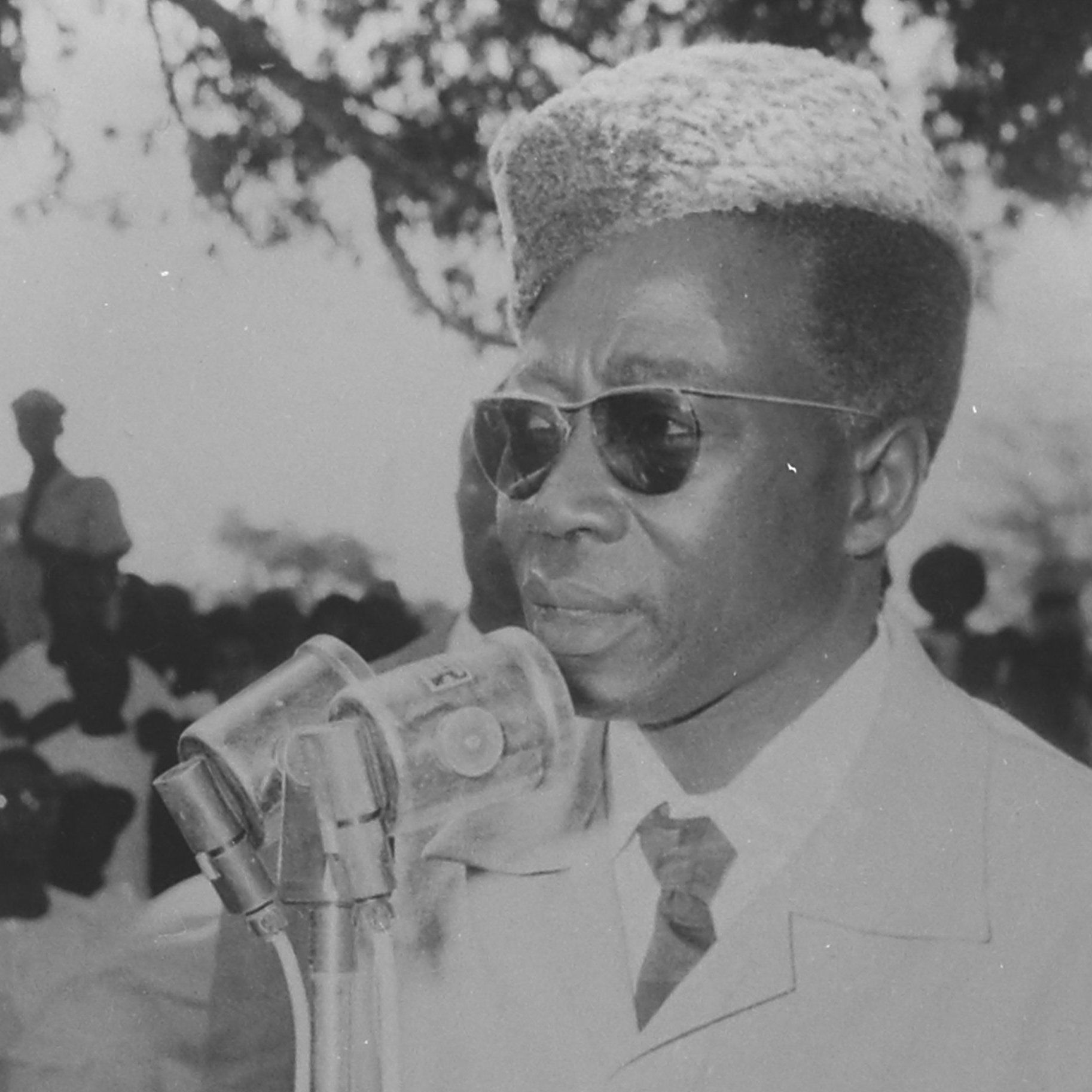 L'image montre un homme debout devant un microphone, s'exprimant clairement. Il porte des lunettes de soleil et a une coiffure distinctive, avec une coupe qui semble bien soignée. Il est vêtu d'un costume clair, ce qui ajoute à son allure sérieuse. En arrière-plan, on peut apercevoir une foule de personnes, dont certaines sont floues, semblant écouter attentivement. L'atmosphère de la scène évoque un moment important, peut-être un discours ou une déclaration publique.