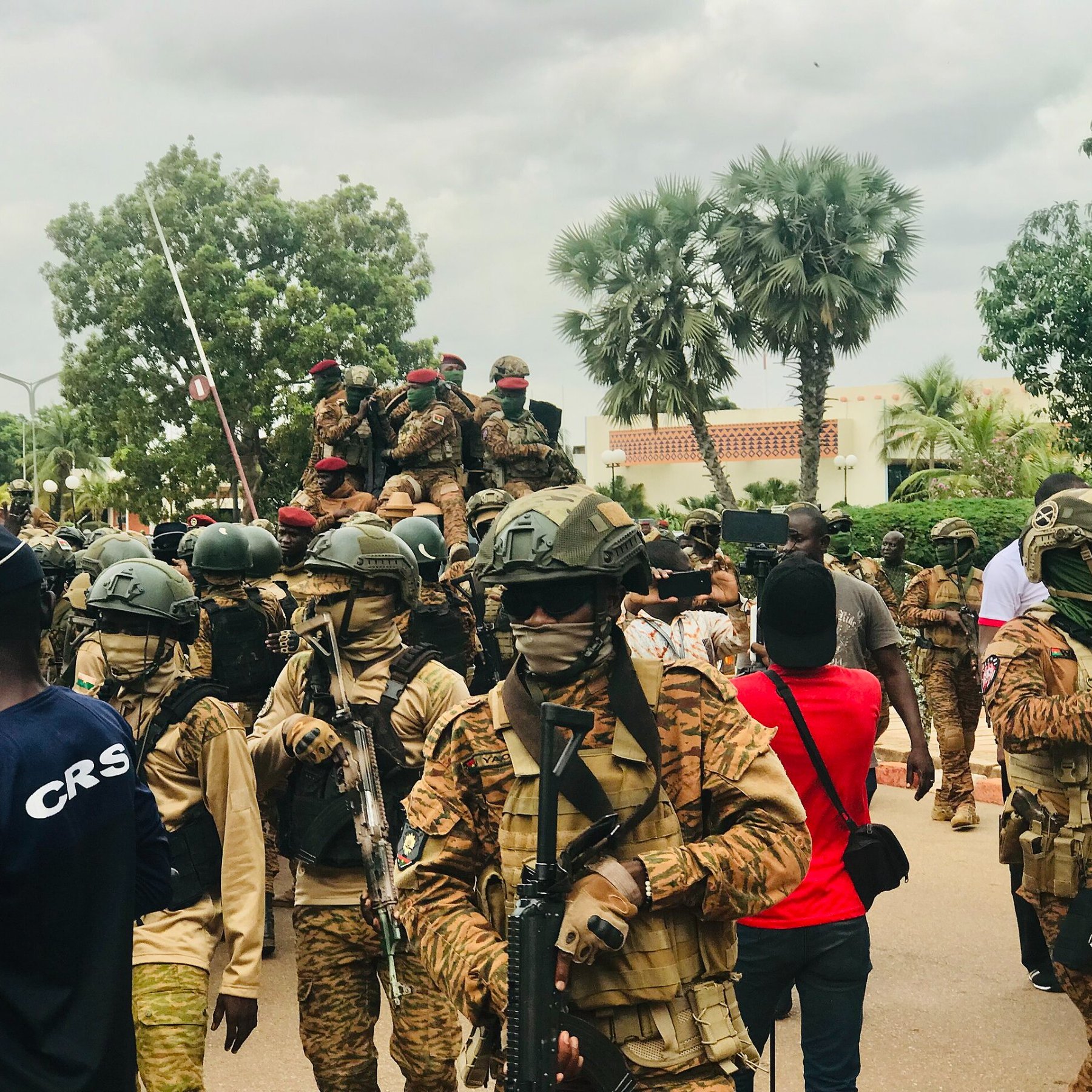 L'image montre une scène animée en plein air, probablement lors d'un événement public ou d'une manifestation. Au premier plan, plusieurs soldats sont regroupés, portant des uniformes militaires et des casques. Ils sont armés et semblent vigilants. L'un d'eux est sur un véhicule militaire, tandis que d'autres avancent sur la route. En arrière-plan, des arbres et des bâtiments peuvent être aperçus, et une foule de citoyens se trouve à proximité, certains portant des vêtements colorés. L'atmosphère peut sembler tendue, surtout en raison de la présence militaire. Le ciel est nuageux, ajoutant une certaine gravité à la scène.