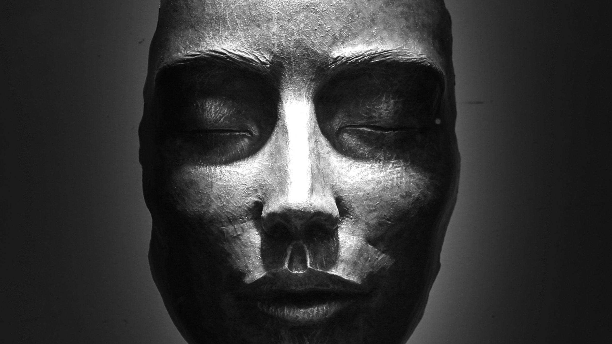L'image représente une sculpture en bronze d'un visage humain. Celui-ci est stylisé et dégage une expression de sérénité. Les yeux sont fermés, et les traits sont à la fois doux et anguleux. La lumière tombe subtilement sur le visage, créant des ombres qui accentuent les contours de la structure. La sculpture est posée sur une base solide, et la posture semble évoquer une sensation de calme et de réflexion. Des détails comme le nez et les lèvres sont finement sculptés, ajoutant une profondeur à l'œuvre. L'ensemble évoque un sentiment de paix intérieure.