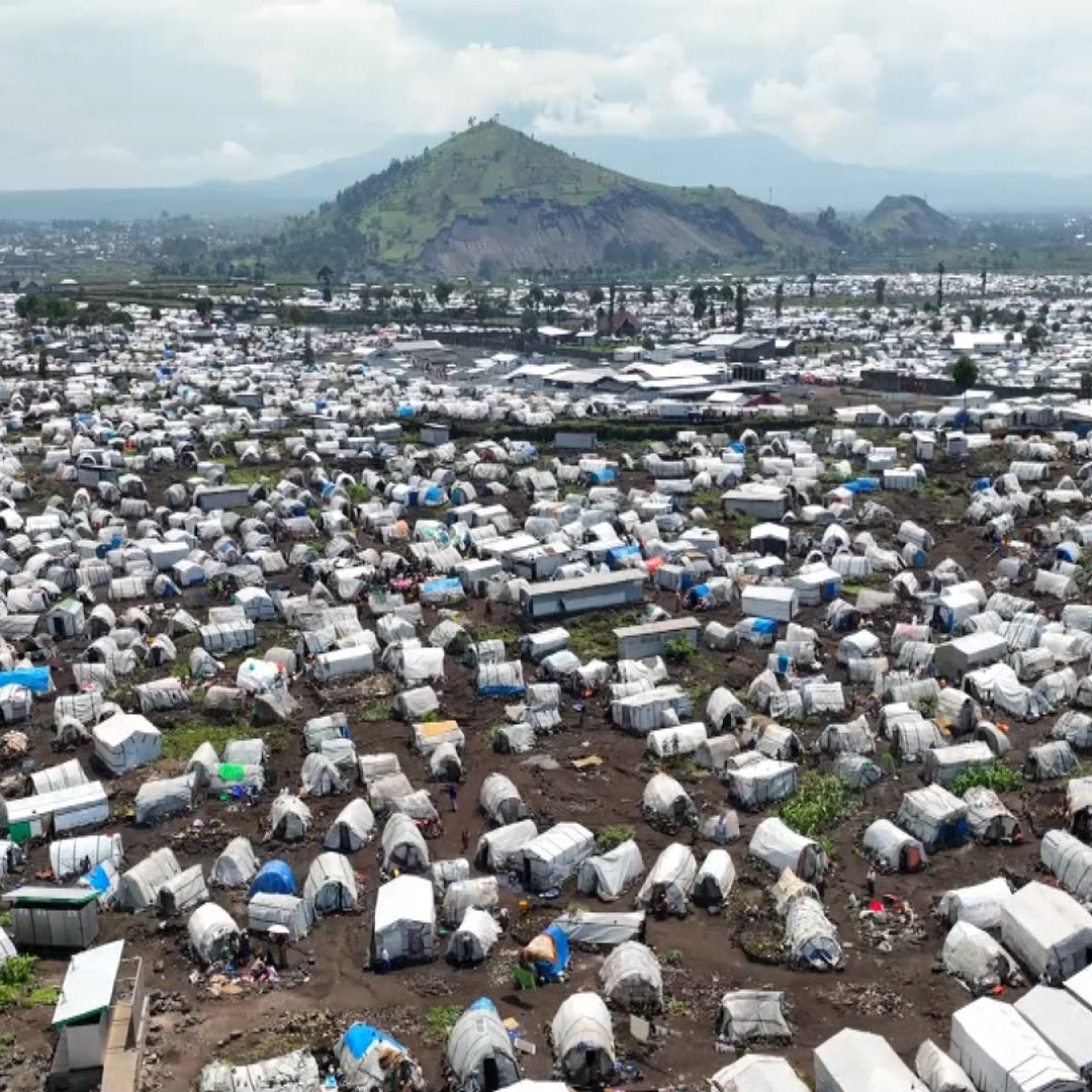 L'image montre un vaste camp de tentes, probablement un site de secours ou un camp de réfugiés, s'étendant à perte de vue. Les tentes sont de couleurs multiples, avec des toits en toile qui varient du blanc au bleu, et certaines sont en désordre avec des morceaux de tissu et de plastique. Le terrain est accidenté, avec des traces d'usage et de la terre visible entre les tentes. En arrière-plan, on aperçoit une montagne verdoyante qui domine le paysage, ajoutant un contraste entre la nature et l'afflux humain. À distance, on distingue des constructions urbaines, des routes et d'autres installations, suggérant la proximité d'une ville. L'ensemble de la scène véhicule une impression de densité et de survie collective, caractéristique des situations d'urgence humanitaire.