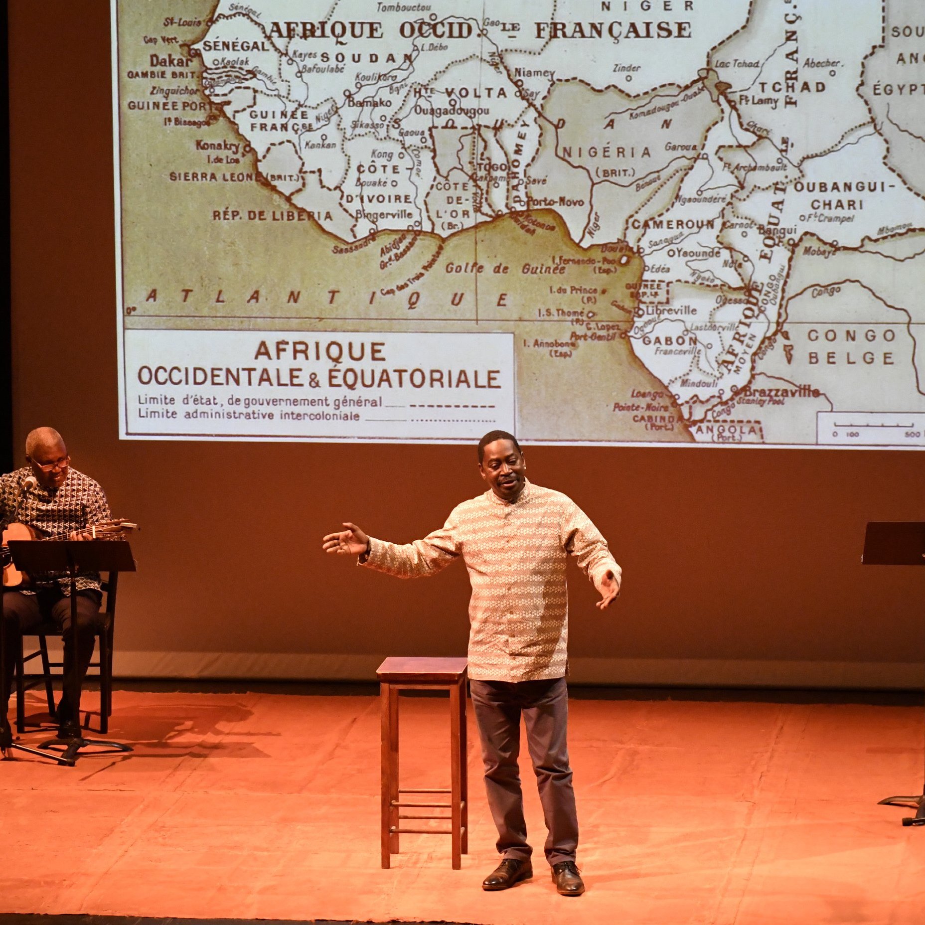L'image représente un spectacle sur scène. Au premier plan, un homme s'exprime avec passion, les bras légèrement écartés pour accentuer son discours. Il porte une chemise claire et semble captivé par son sujet. À sa droite, un autre homme joue de la guitare assis sur un tabouret, ajoutant une ambiance musicale à la performance. En arrière-plan, un grand écran projette une carte ancienne de l'Afrique, spécifiquement d'Afrique occidentale et équatoriale, avec des contours de pays et des indications historiques. Le sol de la scène est décoré de brins de paille, ajoutant une touche naturelle à la mise en scène. L'éclairage doux met en valeur les acteurs et crée une atmosphère intimiste.