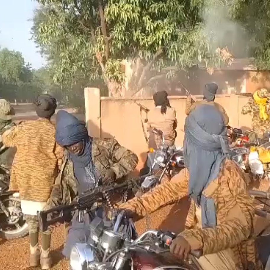 L'image montre un groupe de personnes sur des motos, portant des vêtements qui masquent leur visage et leur tête, probablement pour des raisons de camouflage ou de protection. Ils semblent être dans un environnement extérieur, avec des arbres et un bâtiment en arrière-plan. Il y a de la fumée qui s'échappe, indiquant peut-être une situation de tension ou d'activité intense. Certains individus tiennent des armes, ajoutant une atmosphère de gravité à la scène. Les motos sont variées et leur position suggère qu'ils sont en mouvement ou prêts à partir.