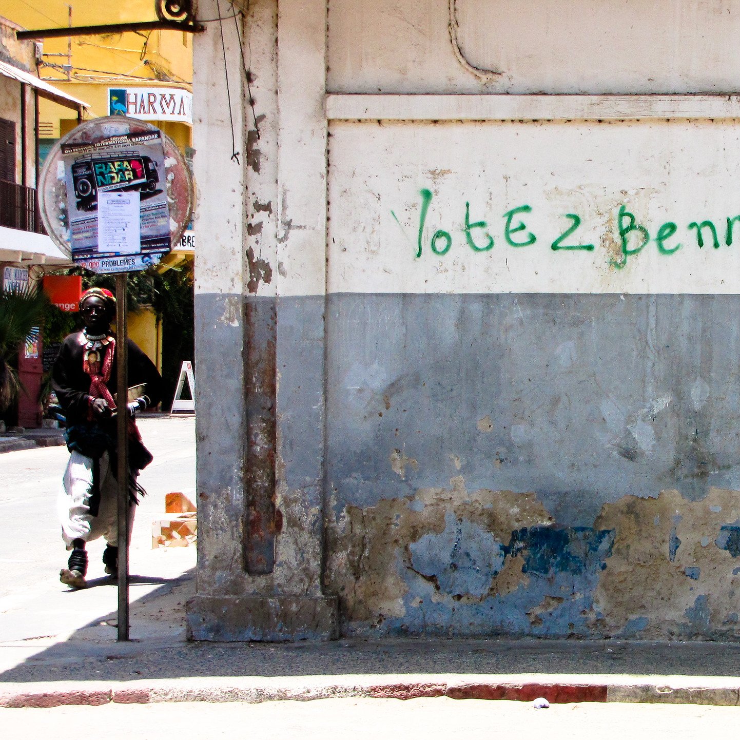 L'image montre un mur d'un bâtiment, principalement de couleur beige, avec des marques d'usure et de décoloration, témoignant de son ancienneté. Une inscription verte, "Votez Benno", est peinte sur le mur. À gauche, on aperçoit la silhouette d'un individu habillé de manière décontractée, qui semble marcher. Au fond, il y a une ambiance animée avec des éléments urbains tels que des affiches et des tables à l'extérieur de cafés. L'ensemble dégage une atmosphère de quartier vivant.