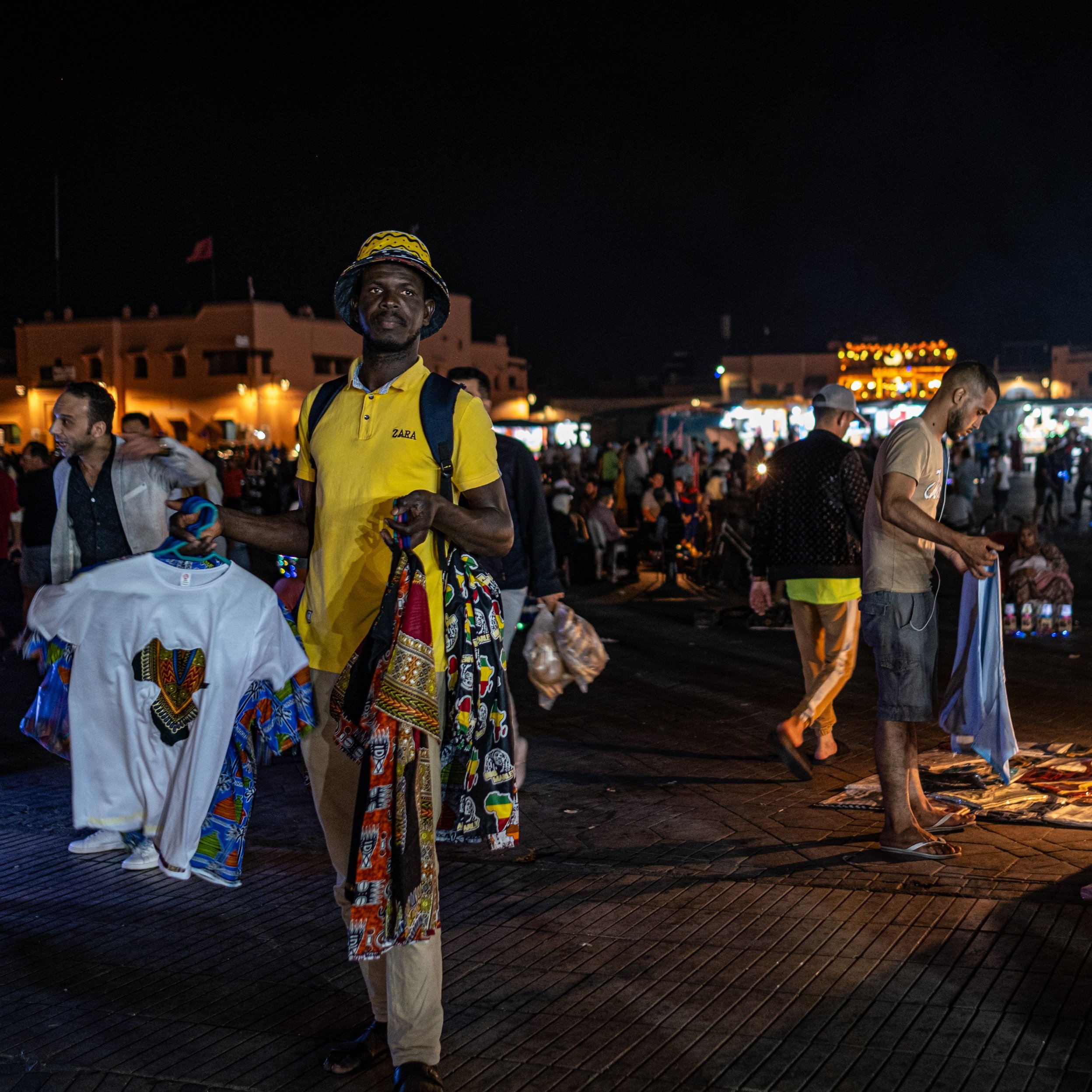 Dans cette image prise la nuit dans un marché animé, on voit un homme au premier plan portant un t-shirt jaune et un chapeau. Il tient plusieurs vêtements, avec des motifs colorés, dans chaque main. L'arrière-plan est rempli de personnes se déplaçant dans le marché, créant une ambiance vivante et dynamique. On peut également apercevoir des lumières brillantes provenant de stands qui éclairent la scène, ajoutant à l'animation nocturne du lieu. La texture du sol semble être en bois ou en pavé, créant un environnement chaleureux. L'air est rempli des sons de conversations et de l'agitation du marché.