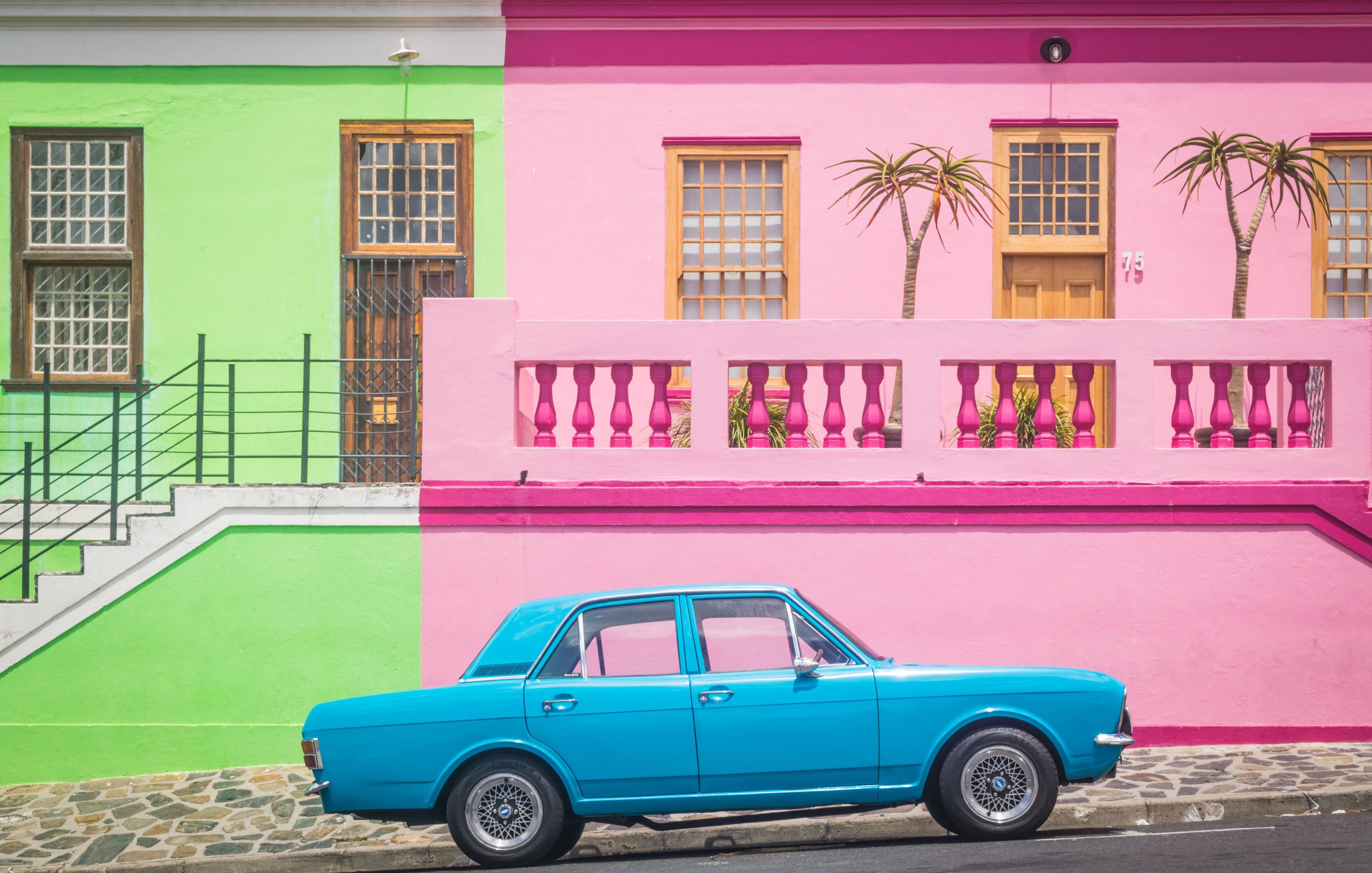L'image présente une scène vibrante et colorée d'une rue typique. À gauche, un bâtiment se dresse en vert clair, avec des fenêtres en bois et un accès par escalier. À droite, un mur rose vif forme la façade d'une maison, ornée de deux palmiers qui ajoutent une touche tropicale. Entre les deux bâtiments, une voiture de couleur bleu clair est garée, se détachant joliment sur le fond coloré. Le sol est pavé, et l'ambiance générale de la scène est joyeuse et accueillante, évoquant une atmosphère estivale et ensoleillée.
