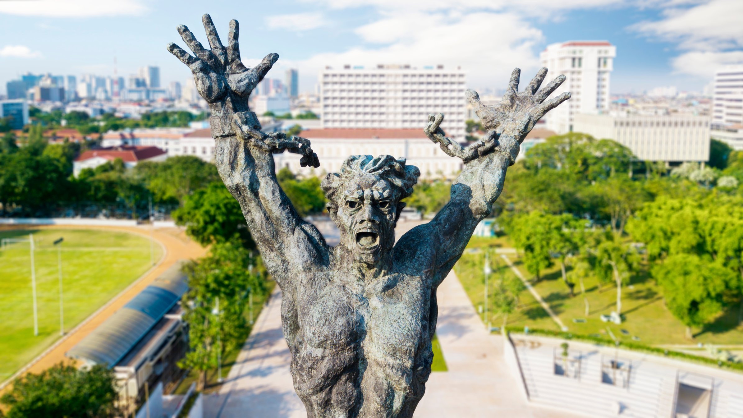 Cette image montre une statue impressionnante d'un homme, représenté dans une pose expressive. Il lève les bras vers le ciel, illustrant un sentiment intense, qui pourrait être celui de la lutte ou de l'angoisse. Son visage est déformé par une expression de détresse, accentuant une impression dramatique. La statue est en bronze, avec une texture riche et des détails finement sculptés qui soulignent les muscles et les traits faciaux. En arrière-plan, on aperçoit une ville moderne avec des bâtiments élevés et des espaces verts, créant un contraste entre la puissance de la sculpture et l'urbanisme environnant. Le ciel est dégagé, ajoutant une luminosité à la scène.