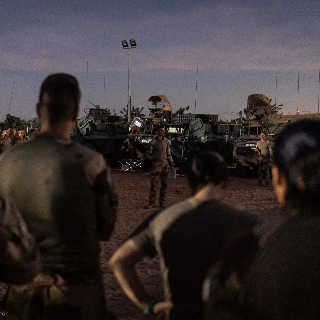 The image depicts a group of military personnel gathered in a semi-dark environment, likely during dusk. They are standing in front of several military vehicles, which appear to be armored and equipped for operational purposes. A leader, possibly giving a briefing or instructions, is positioned in front of the group. The scene conveys a sense of preparation or briefing before a mission or operation. The setting indicates a desert-like terrain, typical for military exercises or deployments in such environments.