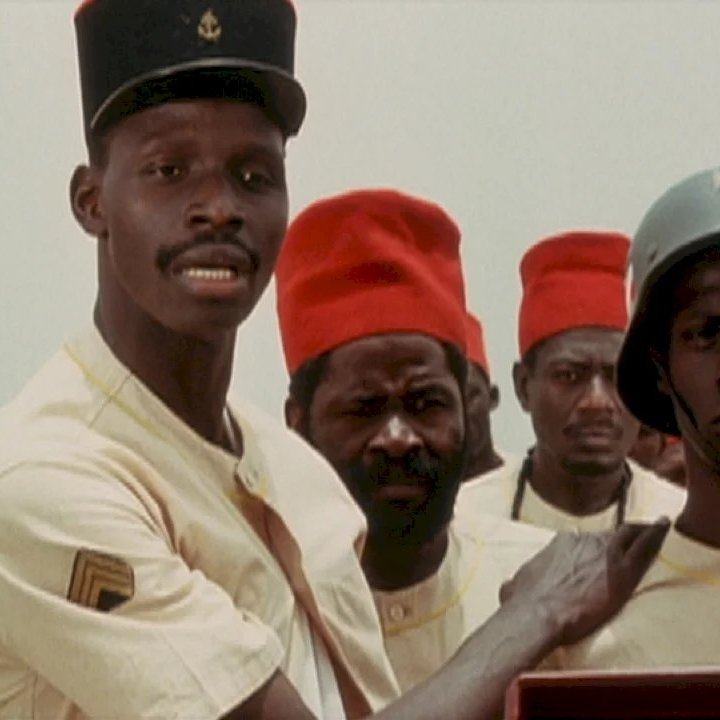 L'image montre plusieurs hommes, en uniformes, rassemblés dans un environnement clair. Au premier plan, un homme se tient à gauche, portant un chapeau noir avec un insigne et une tenue beige. Il regarde vers la droite avec une expression sérieuse. À côté de lui, un autre homme, portant un casque militaire et un uniforme beige, arbore un air grave. Derrière eux, plusieurs autres hommes regardent également dans la même direction. Tous semblent engagés dans une discussion ou une situation importante. Les couleurs dominantes de l'image sont le beige des uniformes et le rouge vif des chapeaux de certains des hommes.