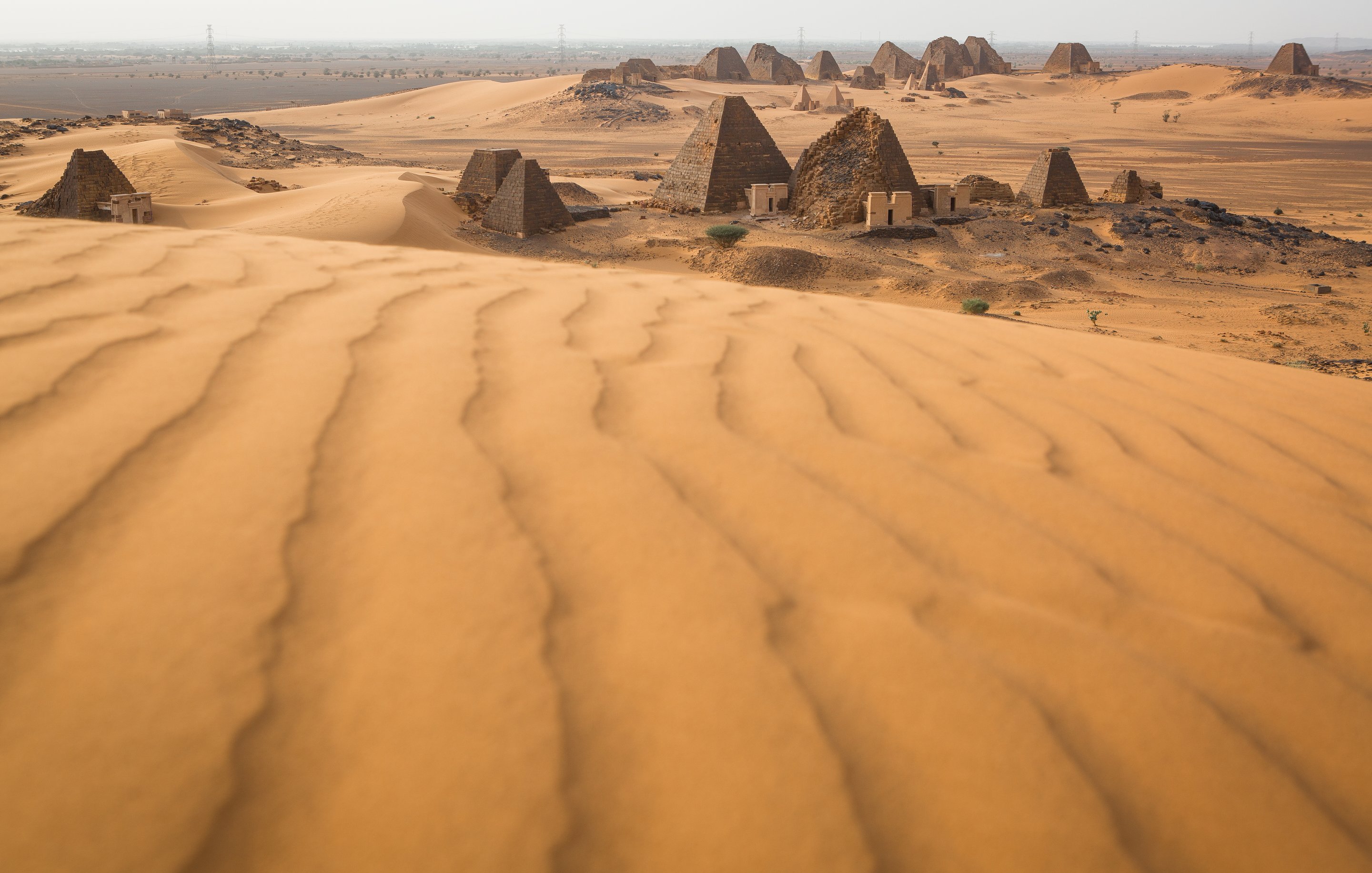 L'image décrit un paysage désertique, dominé par de vastes dunes de sable aux formes ondulantes. En arrière-plan, on aperçoit des pyramides anciennes dont les sommets sont pointus et les surfaces sont qui semblent rugueuses. Le terrain autour des pyramides est également parsemé de petites éboulis et de dunes, créant un paysage à la fois majestueux et mystérieux. La lumière du soleil éclaire la scène, mettant en valeur les nuances dorées du sable et l'architecture historique des pyramides. L'atmosphère générale évoque la chaleur et l'immensité du désert, ainsi qu'un sentiment d'émerveillement face aux vestiges du passé.