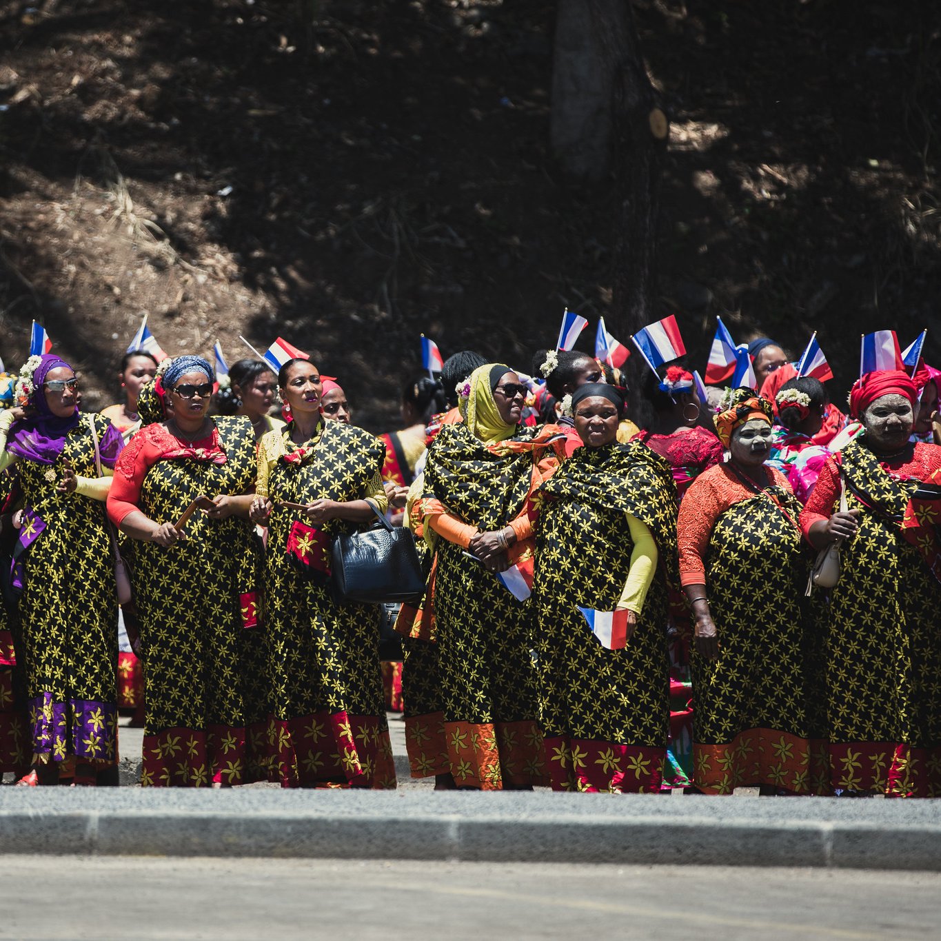 L'image montre un groupe de femmes réunies en ligne, vêtues de robes longues aux motifs floraux colorés. Chaque femme porte des accessoires traditionnels, comme des foulards vifs. Certaines tiennent des drapeaux aux couleurs bleu, blanc et rouge, symbolisant leur fierté nationale. L'arrière-plan est naturel, avec des arbres, et l'ambiance semble festive et engagée, suggérant une célébration ou un événement culturel. Les femmes affichent des expressions de joie et d'unité.