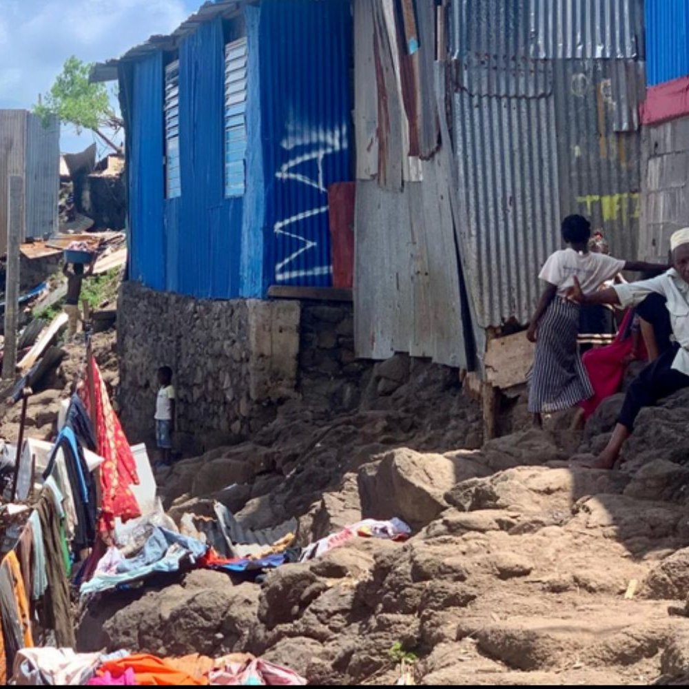 Cette image présente un paysage d'un quartier informel, caractérisé par des habitations en tôle et en béton, construite sur un terrain escarpé. Les maisons, souvent de différentes couleurs, semblent inachevées avec des parois en métal ondulé. Au premier plan, des personnes sont assises sur des rochers, discutant ou s'affairant. Le sol est accidenté, recouvert de débris et de vêtements épars, tandis qu'en arrière-plan, des structures réduites à des vestiges montrent l'impact de l'environnement sur ce lieu de vie. Le ciel est clair, laissant présager une journée ensoleillée. L'atmosphère générale évoque une communauté résiliente, malgré les difficultés et les conditions de vie précaires.