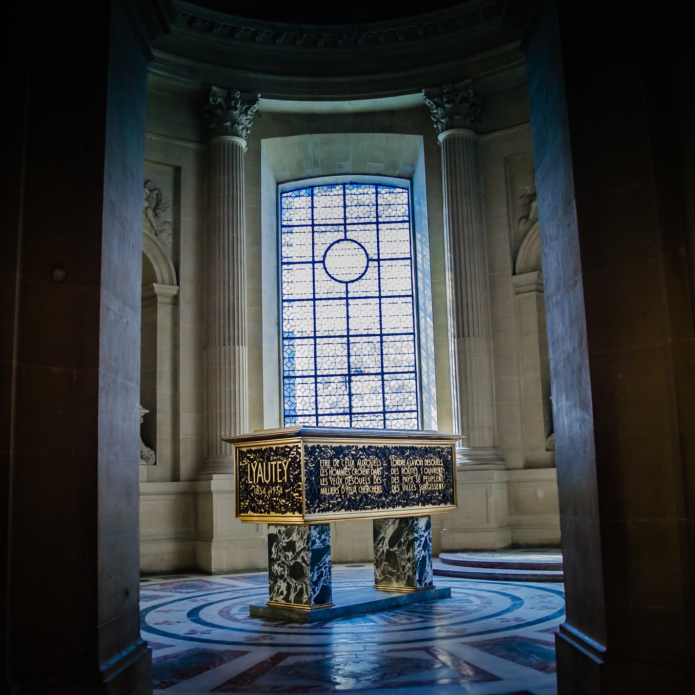 Cette image montre une pièce d'un bâtiment majestueux, probablement un musée ou un palais. Au centre, se trouve un coffre sombre et décoré, surélevé sur un socle circulaire. Les côtés du coffre sont ornés de motifs complexes et d'inscriptions dorées. En arrière-plan, une grande fenêtre ornée de vitraux diffuse une lumière douce qui éclaire la pièce. Les murs sont en pierre grise, enrichis de colonnes majestueuses. L'atmosphère générale est à la fois calme et solennelle, invitant à la contemplation.