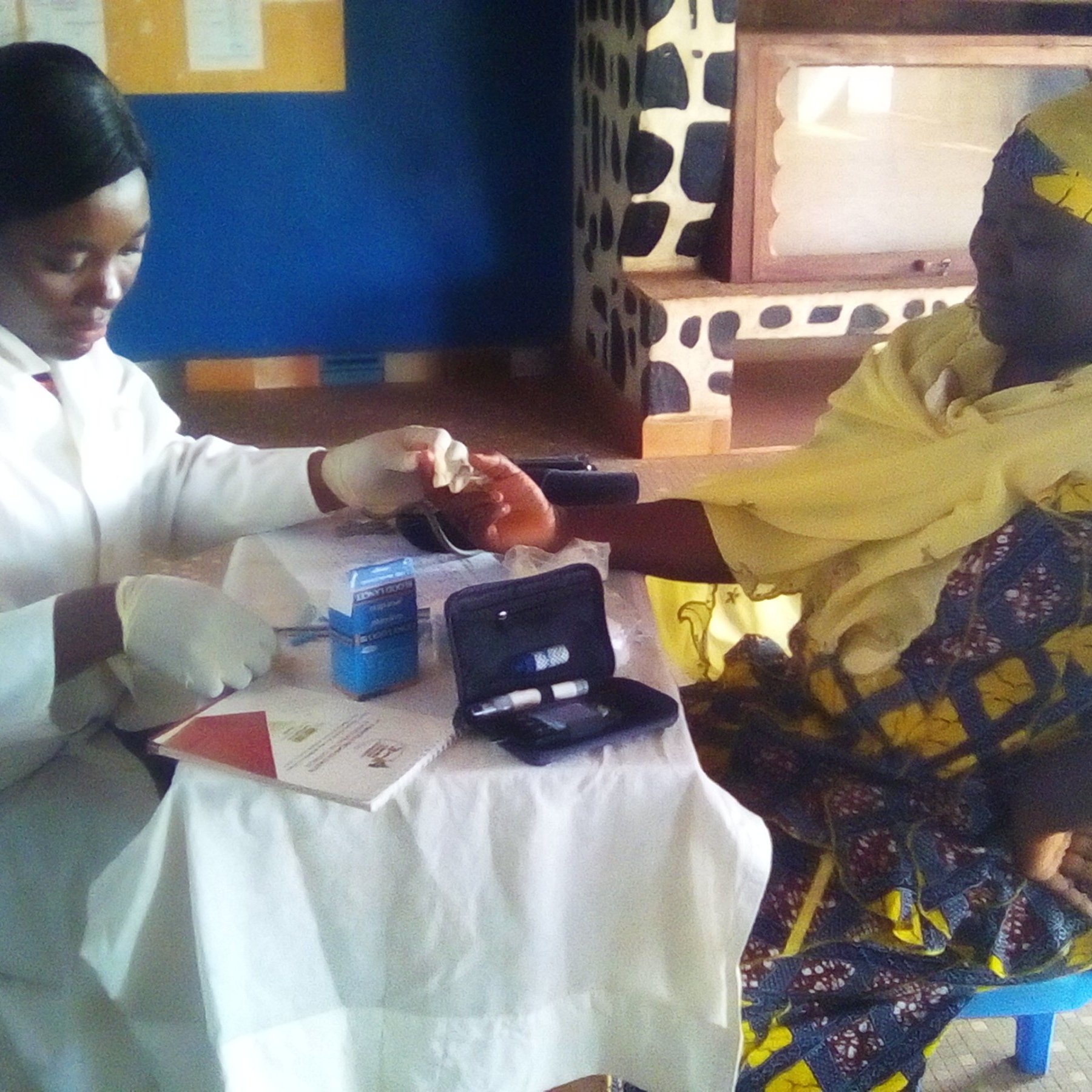 L'image montre une scène dans une salle de consultation. À gauche, une professionnelle de la santé, vêtue d'un manteau blanc et de gants, effectue un test sur le bras d'une patiente. La femme qui reçoit le soin porte un turban jaune et une robe aux motifs colorés. Elle semble concentrée et un peu nerveuse. Sur la table devant elles, on peut voir des documents et des instruments médicaux, comme un petit appareil de mesure. Les murs de la pièce sont peints en bleu, et le sol est en béton. L'environnement est calme, et l'atmosphère est professionnelle, axée sur le soin et le soutien.