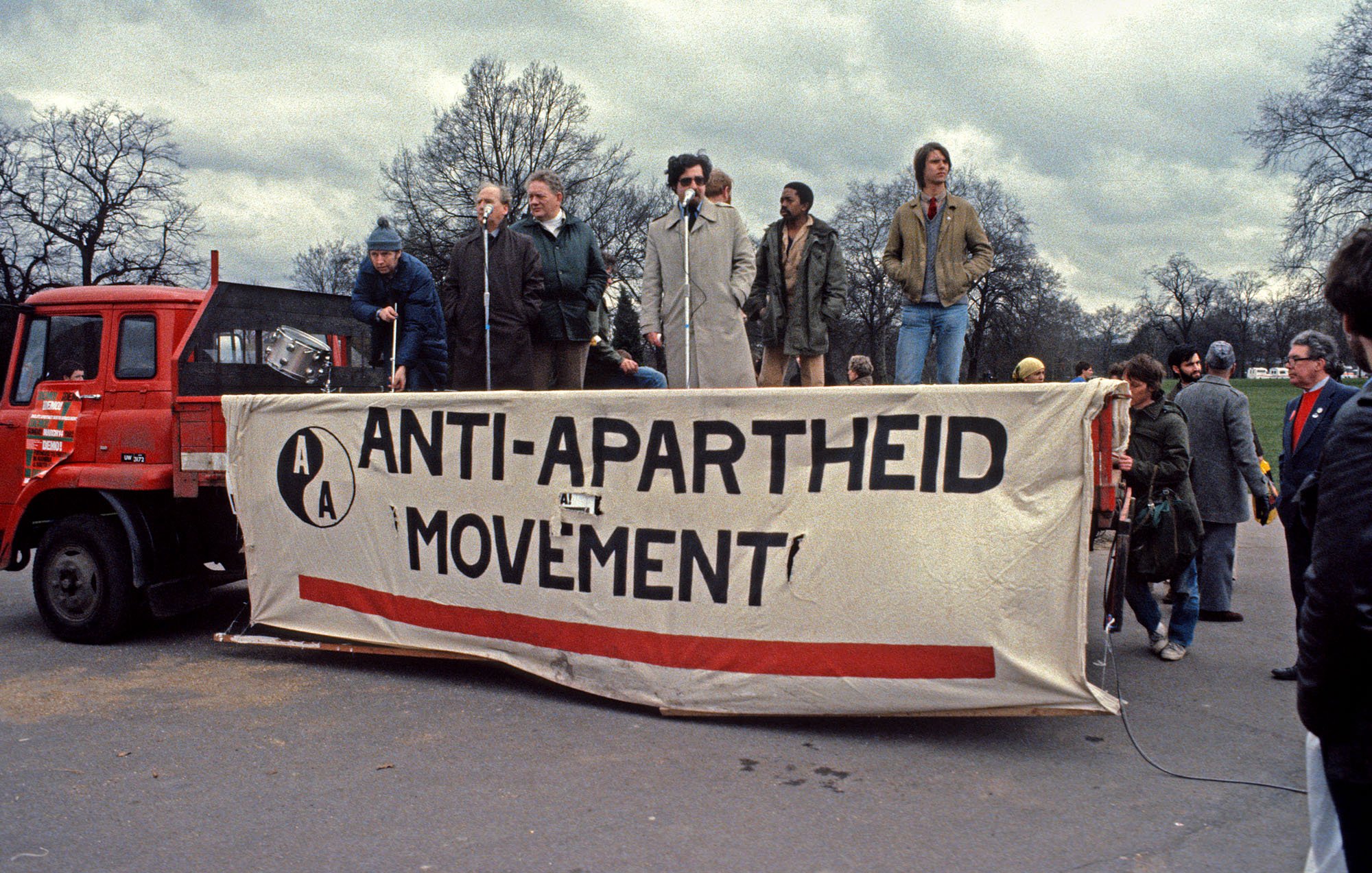 Cette image représente un rassemblement pour le mouvement anti-apartheid. On voit une scène sur un camion, avec plusieurs personnes qui parlent à un microphone. La bannière au premier plan affiche le texte "ANTI-APARTHEID MOVEMENT" en lettres majuscules, souligné par une bande rouge. L'environnement est urbain, avec des arbres dénudés en arrière-plan et un ciel nuageux, ce qui crée une atmosphère sérieuse et engagée. Les participants semblent passionnés, témoignant de l'importance de la lutte pour l'égalité et les droits civiques.
