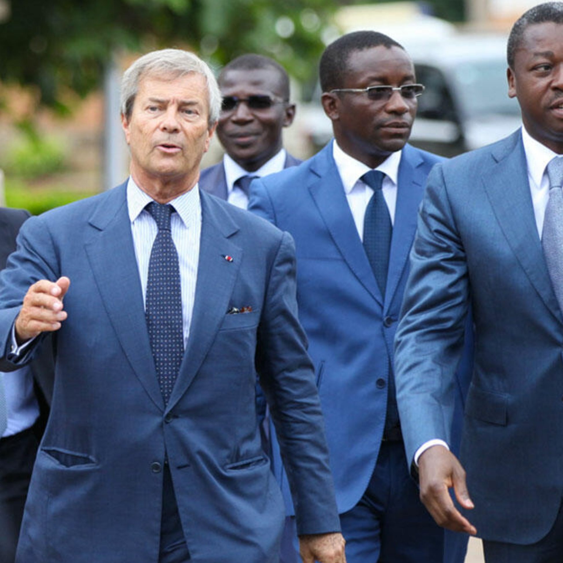 L'image montre un groupe d'hommes marchant ensemble. Le premier homme, à gauche, porte un costume bleu clair et une cravate à rayures. Il semble engagé dans la conversation. Le deuxième homme, au centre, porte aussi un costume bleu, avec une chemise blanche. Il a un regard sérieux et écoute attentivement. Le troisième homme, à droite, est vêtu d'un costume bleu foncé et marche avec assurance, affichant un léger sourire. En arrière-plan, on aperçoit d'autres individus et un environnement qui suggère un événement ou une rencontre formelle. Les hommes semblent dans un contexte professionnel.