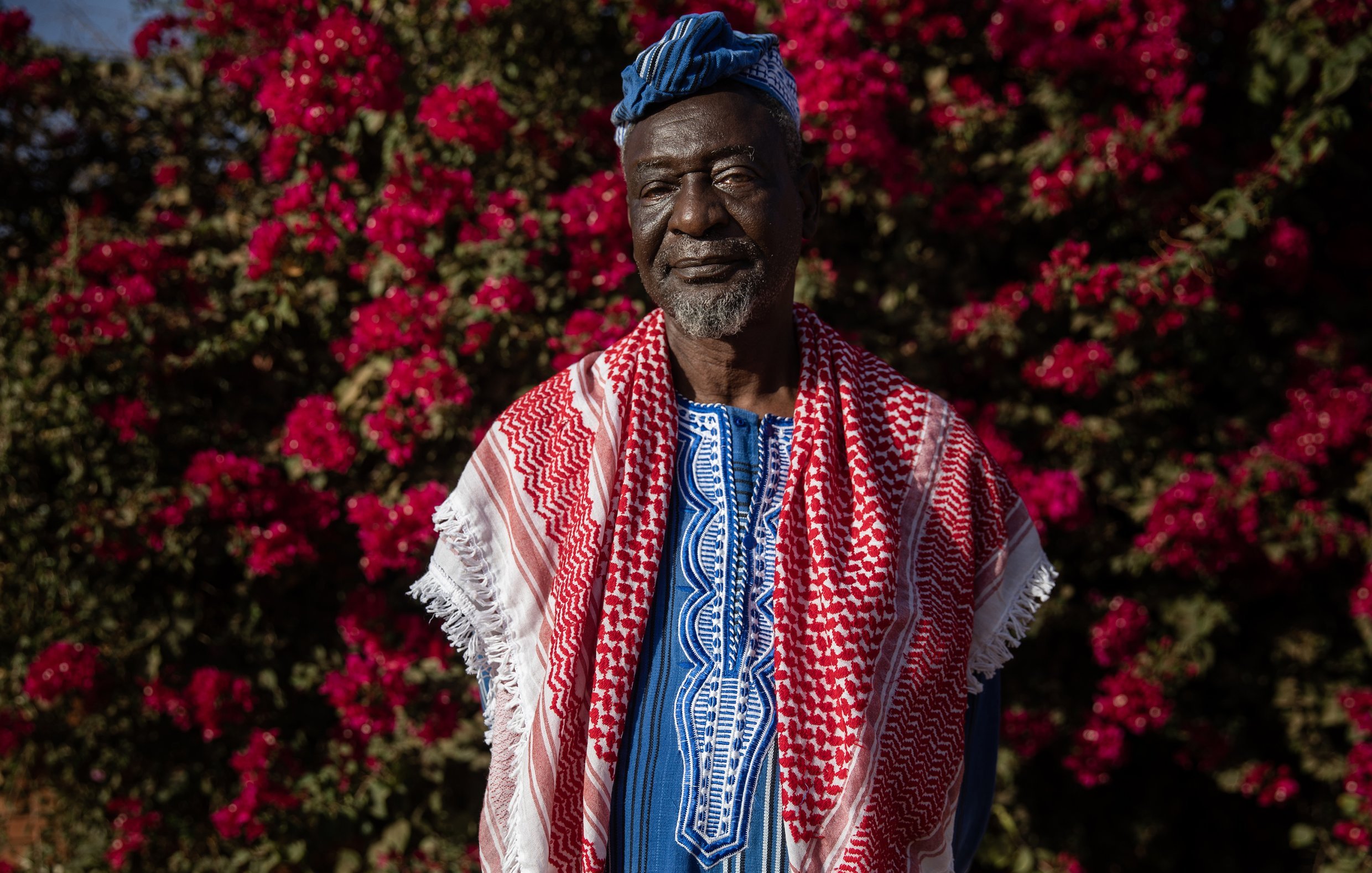 L'image montre un homme debout devant un fond vibrant de fleurs roses, probablement des bougainvilliers. Il porte une tenue traditionnelle riche en couleurs, avec un tissu bleu et des motifs complexes. Sur ses épaules, il a une écharpe ou un châle rouge et blanc, typique de certaines cultures. Son visage exprime sagesse et dignité, avec des traits marqués par le temps. Le contraste entre ses vêtements et les fleurs éclatantes crée une atmosphère chaleureuse et accueillante.