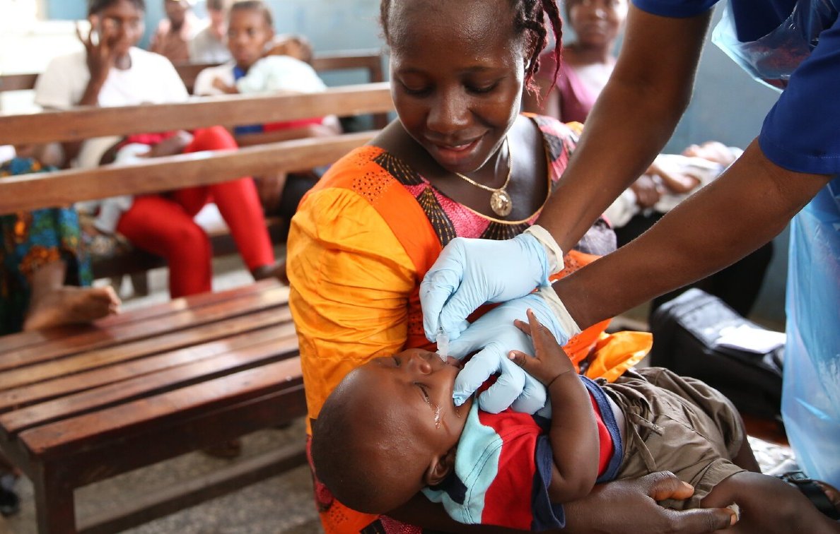 Dans cette image, nous voyons une scène dans une salle d'attente où plusieurs personnes sont assises sur des bancs en bois. Au premier plan, une mère, vêtue d'une robe orange, tient son bébé. Elle semble attentive et douce. Un professionnel de la santé, portant des gants bleus, est en train de donner un soin ou un médicament au bébé. Le bébé, habillé d'un t-shirt bleu et d'un short, a une expression calme. À l'arrière-plan, d'autres femmes et enfants attendent, créant une atmosphère de communauté et de soutien. Les murs sont peints en bleu, ajoutant une ambiance sereine à la scène.