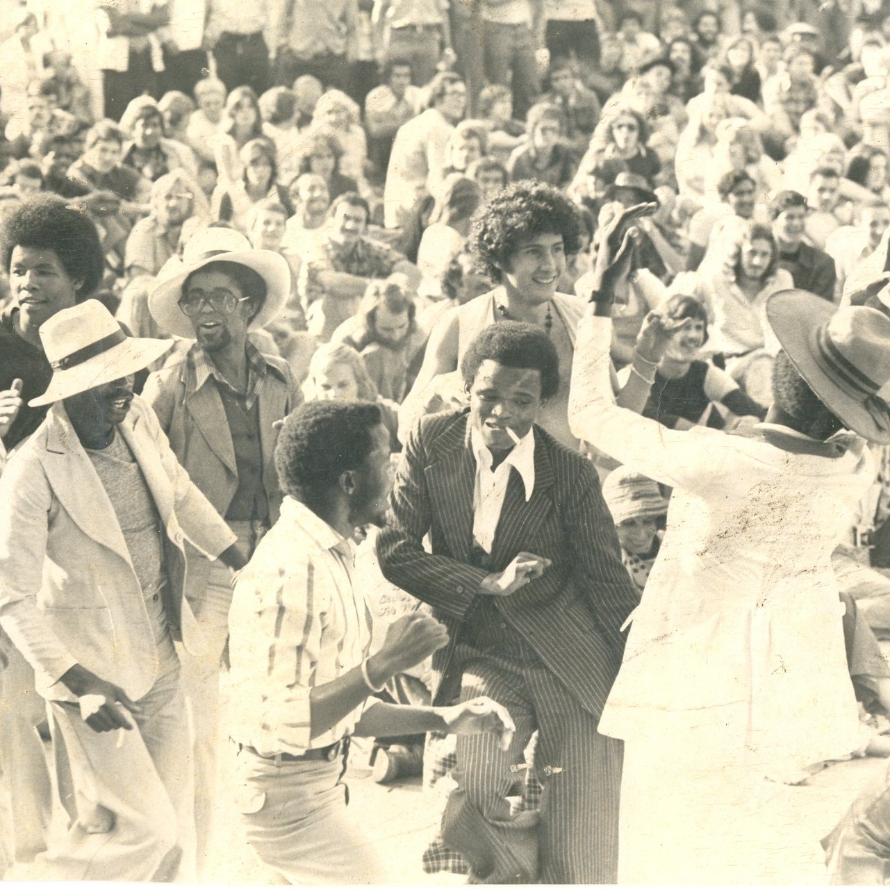 L'image représente une scène vibrante de fête en plein air, probablement lors d'un concert ou d'un festival. Au premier plan, un groupe de personnes danse joyeusement, exprimant de l'énergie et de la convivialité. Les danseurs, de différentes origines, portent des vêtements variés, allant des costumes élégants à des tenues plus décontractées. En arrière-plan, une foule dense assiste à la scène, créant une ambiance de camaraderie. Les visages sont souriants, captivés par la musique et l'atmosphère festive. Le tout suggère une célébration de la vie et de la culture, où la danse et la communion entre les gens sont à l'honneur. L'éclairage semble naturel, évoquant une belle journée ensoleillée.