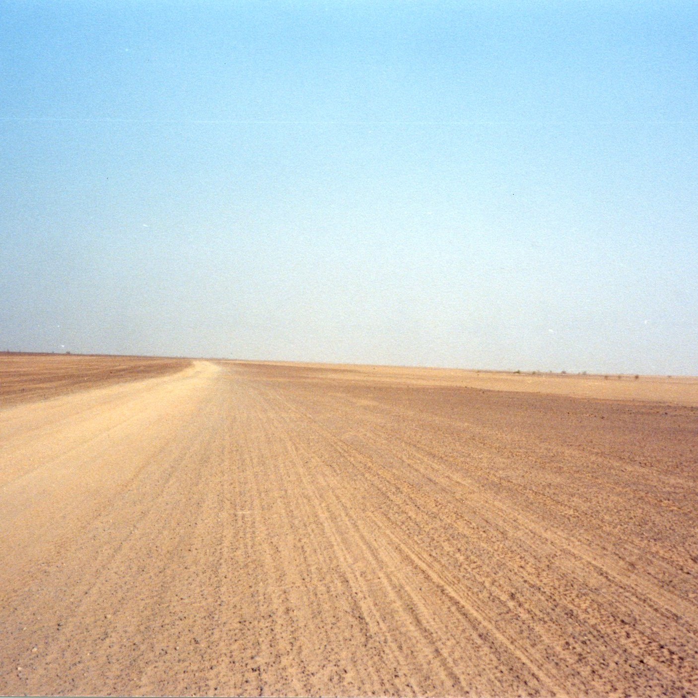 Imaginez un vaste paysage désertique, s'étendant à perte de vue sous un ciel bleu clair. Le sol est constitué de sable et de terre, créant une texture rugueuse sous vos pieds. La route est légèrement sinueuse, formant des lignes qui se fondent avec l'horizon. À première vue, tout semble calme et silencieux, avec peu de perturbations, excepté pour le vent. Sur les bords de ce paysage, on peut percevoir des nuances jaune et beige, typiques des déserts. L'atmosphère est sereine, presque méditative, laissant une impression de solitude face à l'immensité de la nature.