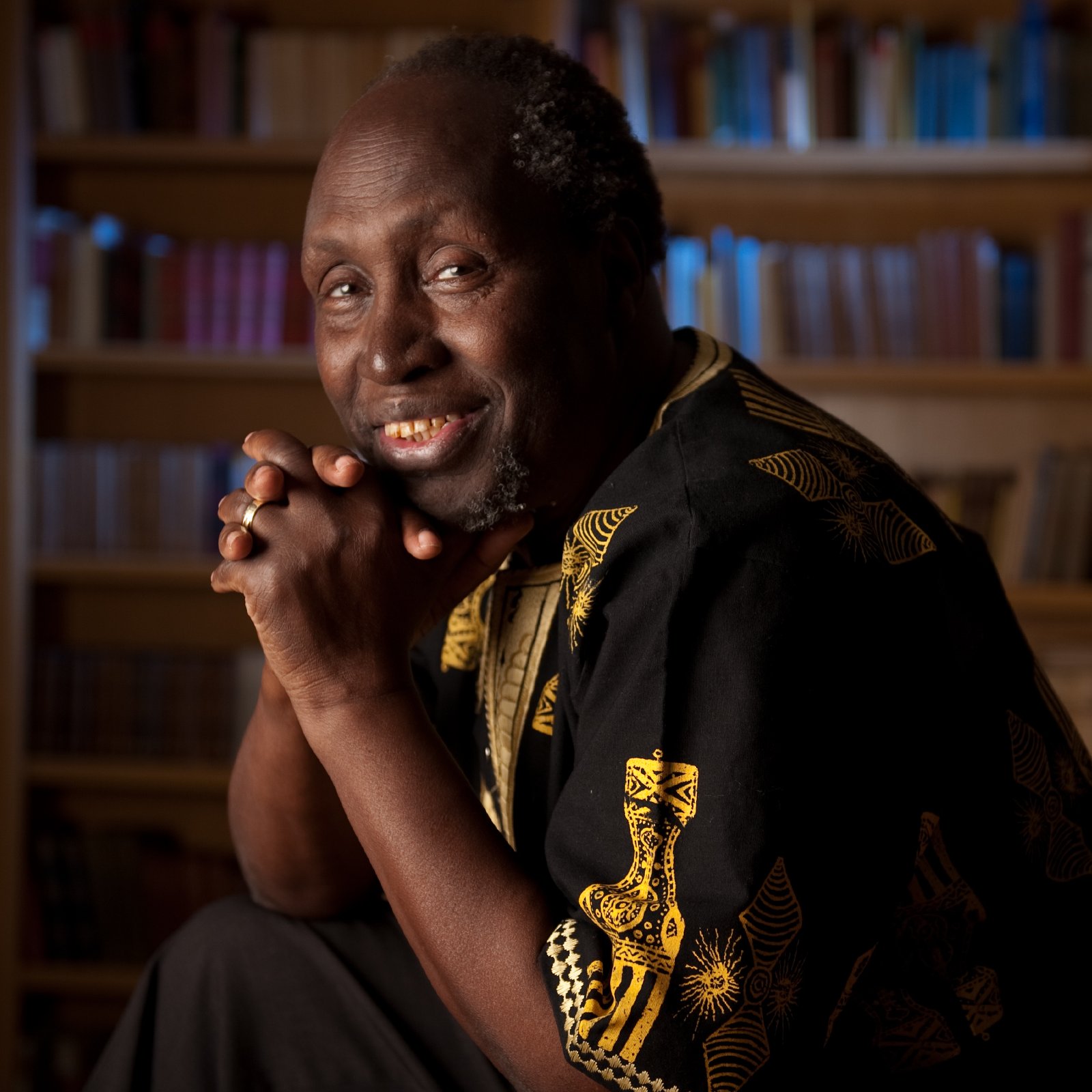 L'image montre un homme assis dans une bibliothèque, entouré de nombreuses étagères remplies de livres. Il a un sourire chaleureux et une expression amicale. Il porte une chemise noire ornée de motifs dorés, ce qui ajoute une touche d'élégance à son apparence. Son visage exprime une sagesse et une bienveillance, tandis qu'il a les mains croisées sous son menton. L'arrière-plan est légèrement flou, mettant en valeur sa figure et créant une atmosphère accueillante et studieuse.