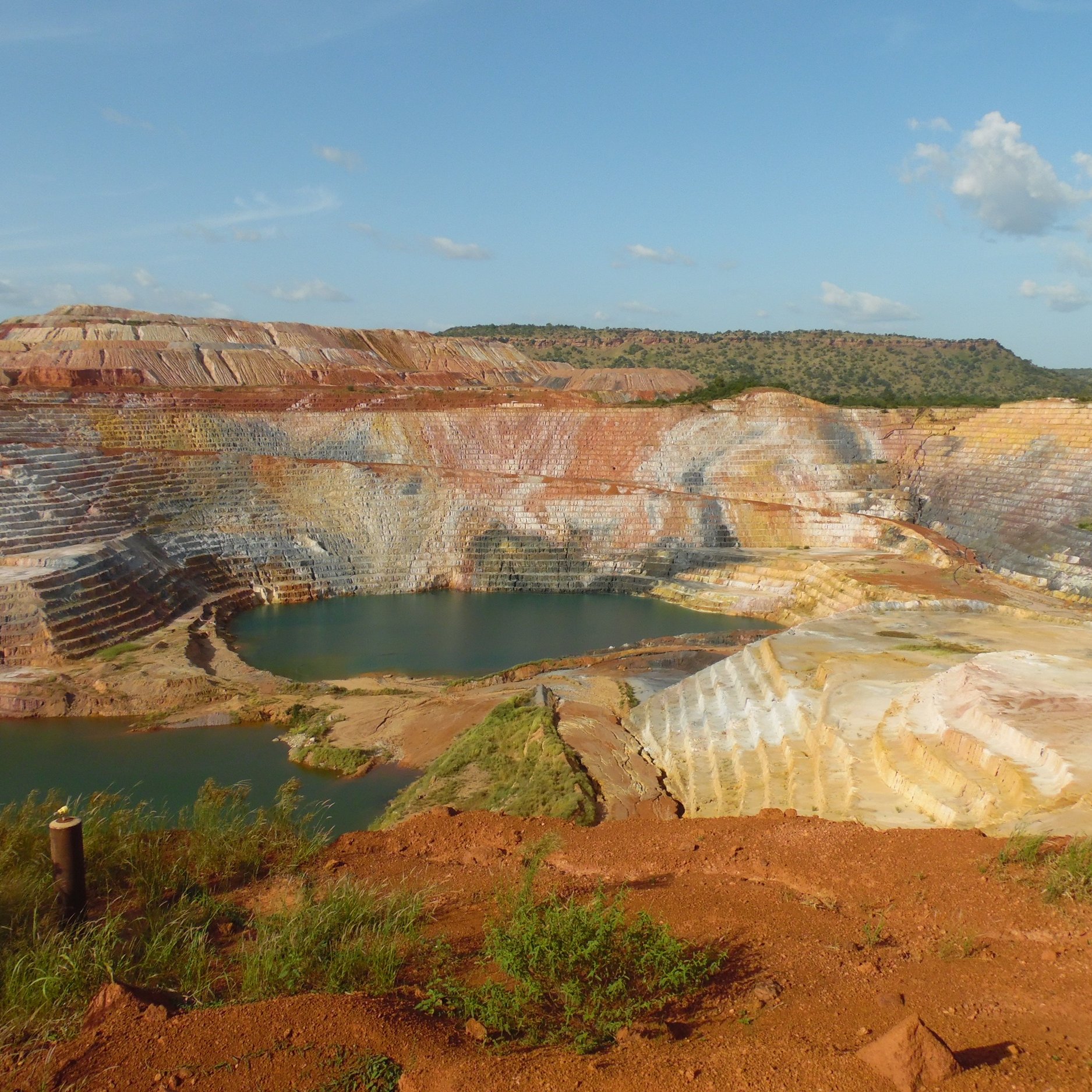 L'image montre une carrière de minerai, un paysage dramatique où les strates de terre créent des dégradés de couleurs allant du rouge au jaune et au blanc. Les parois de la carrière sont escarpées, formant des niveaux bien distincts. Au centre, un plan d'eau bleu émerge, contrastant avec les teintes terreuses et les paysages environnants. La végétation se fait rare sur les bords, mais quelques petites herbes vertes remontent le long des pentes. Le ciel est clair avec quelques nuages épars, ajoutant une lumière douce à la scène. Cette vue évoque à la fois l'exploitation humaine et la beauté naturelle brute.