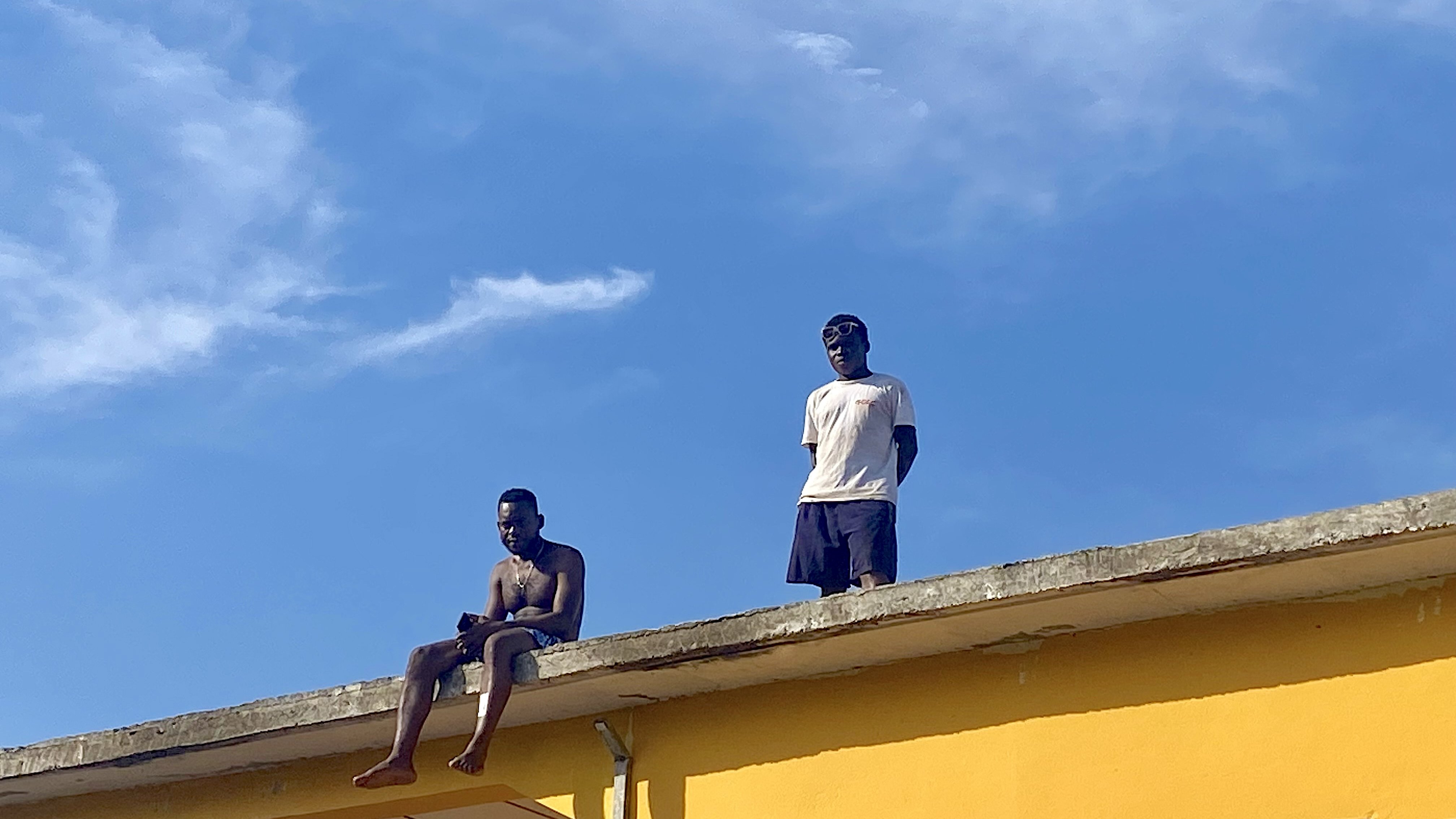 Sur cette image, deux jeunes hommes se trouvent sur le toit d'une maison peinte en jaune vif. L'un d'eux est assis au bord, les jambes pendantes dans le vide, tandis que l'autre se tient debout à ses côtés. Le ciel est clair et bleu, avec quelques nuages légers. L'ambiance semble détendue, peut-être un moment de loisirs ou de camaraderie. Les détails de l'environnement autour d'eux ne sont pas visibles dans cette capture, mais l'accent est mis sur leur posture et l'interaction entre eux.