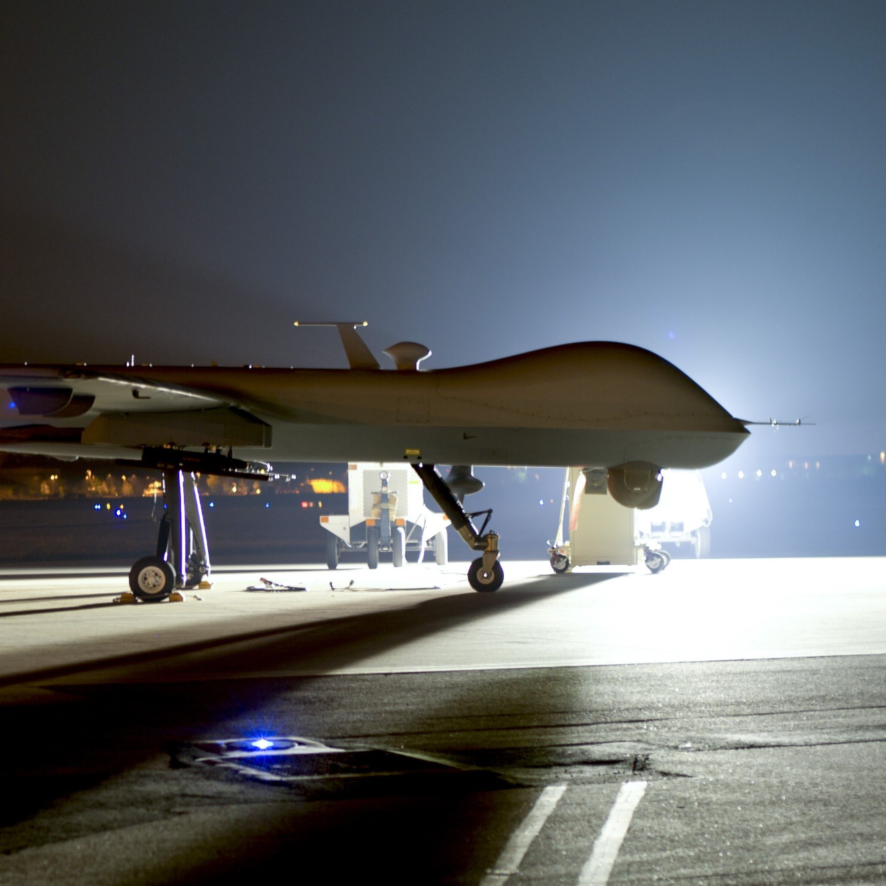 L'image montre un drone de surveillance, typiquement de type militaire, stationné sur une piste d'atterrissage. La scène est nocturne, éclairée principalement par des lumières artificielles qui révèlent les contours du drone. Le drone a une forme allongée et est équipé d'une hélice à l'arrière. Il repose sur un châssis avec des supports, et il y a des lumières au sol qui peuvent délimiter la zone ou indiquer l'emplacement. L'ambiance est sérieuse et technologique, évoquant des missions de surveillance ou de reconnaissance.
