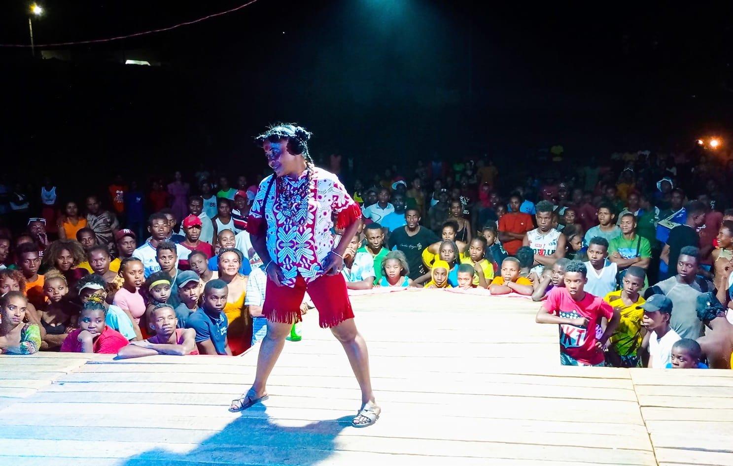 L'image montre une scène vivante et animée. Au premier plan, un artiste, habillé de manière colorée avec une chemise à motifs et un short, se tient sur une scène en bois. Son expression enthousiaste et son attitude dynamique suggèrent qu'il est en plein spectacle. Derrière lui, une foule nombreuse est présente, composée de personnes de tous âges et de toutes origines, certaines souriant et d'autres captivées par la performance. Les lumières autour de la scène créent une atmosphère festive, tandis que l'obscurité en arrière-plan met l'accent sur l'énergie du moment. Les spectateurs semblent engager et réceptifs, témoignant de l'ambiance joyeuse d'un événement en plein air.