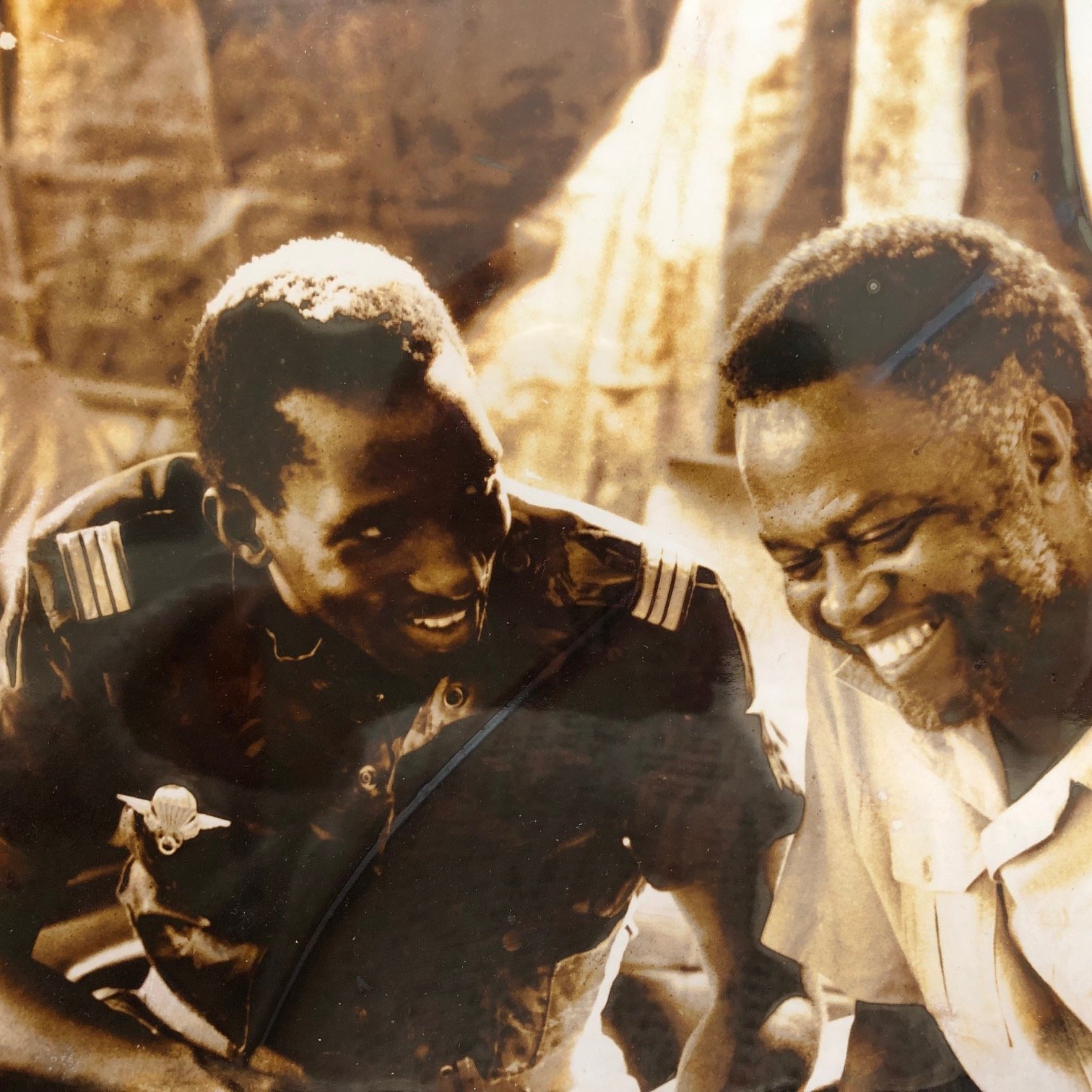 L'image montre deux hommes assis côte à côte, visiblement en train de discuter et de partager un moment de complicité. L'un des hommes porte un uniforme militaire, tandis que l'autre est vêtu d'une chemise claire. Ils affichent des sourires sincères, ce qui dénote une ambiance chaleureuse et amicale. L'arrière-plan est flou, mais on peut deviner une scène informelle, peut-être un rassemblement ou une réunion. La photo donne une impression de camaraderie et de joie partagée entre les deux individus.