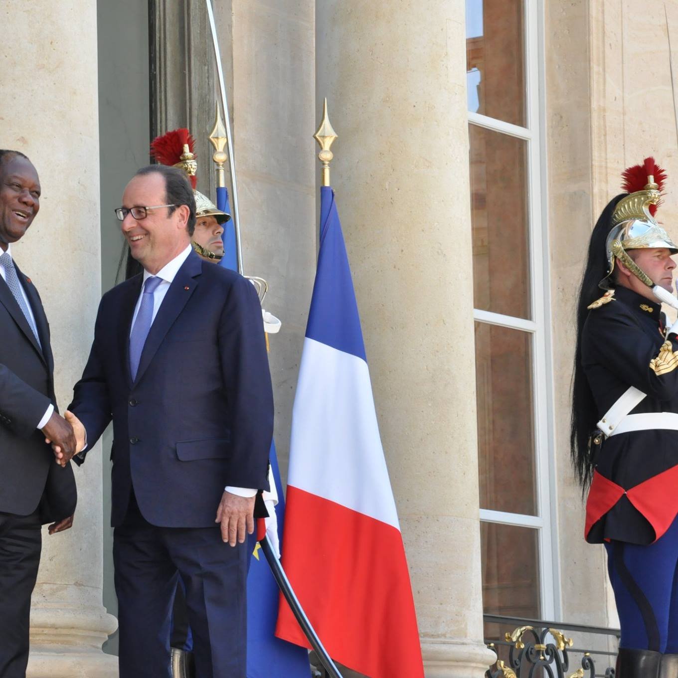 The image shows two men shaking hands outside a large building, likely a government or official residence. One man is wearing a dark suit and the other is dressed in a suit and tie, likely representing different nations. Behind them, there is a large flag of France, and guards in traditional uniforms stand watch. The setting suggests a formal meeting or diplomatic engagement.
