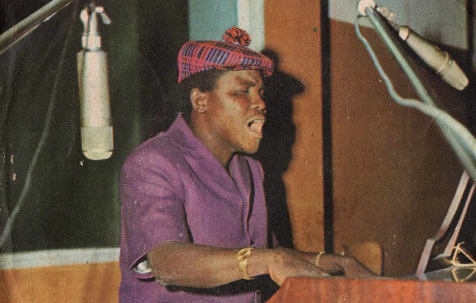 Sur cette image, nous voyons un homme en train de jouer du piano. Il porte une chemise violette et un chapeau à motif écossais. Son expression est concentrée et passionnée alors qu'il chante. Deux microphones sont placés à proximité, capturant sa voix. L'arrière-plan présente des panneaux en bois colorés, créant une ambiance chaleureuse et artistique. L'atmosphère dégage une sensation de musique vivante et d'émotion.