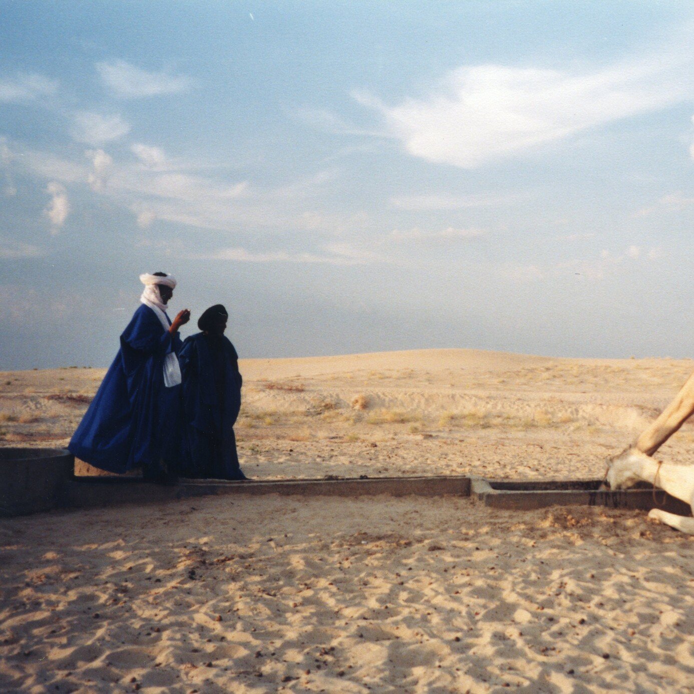 L'image montre un paysage désertique, caractérisé par des dunes de sable qui s'étendent à perte de vue sous un ciel légèrement nuageux. Au premier plan, deux personnes, habillées de vêtements traditionnels, se tiennent près d'un point d'eau. L'une d'elles, en bleu, semble prier ou méditer. À proximité, un chameau repose tranquillement sur le sol, tandis qu'un autre chameau se penche pour boire. L'ambiance de la scène est paisible, avec une lumière douce qui souligne les formes des dunes et des silhouettes. Les sons du désert, comme le vent léger, pourraient renforcer cette atmosphère sereine.