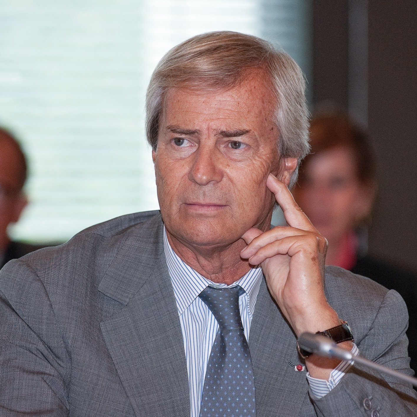 L'image montre un homme d'âge moyen, assis à une table de conférence. Il a les cheveux gris et porte un costume gris. Son expression est pensive, et il touche son visage avec un doigt en réfléchissant. En arrière-plan, on peut distinguer d'autres personnes assises, mais leurs visages sont flous. L'ambiance semble professionnelle, suggérant une réunion ou une discussion importante. La lumière est douce, créant une atmosphère de concentration.