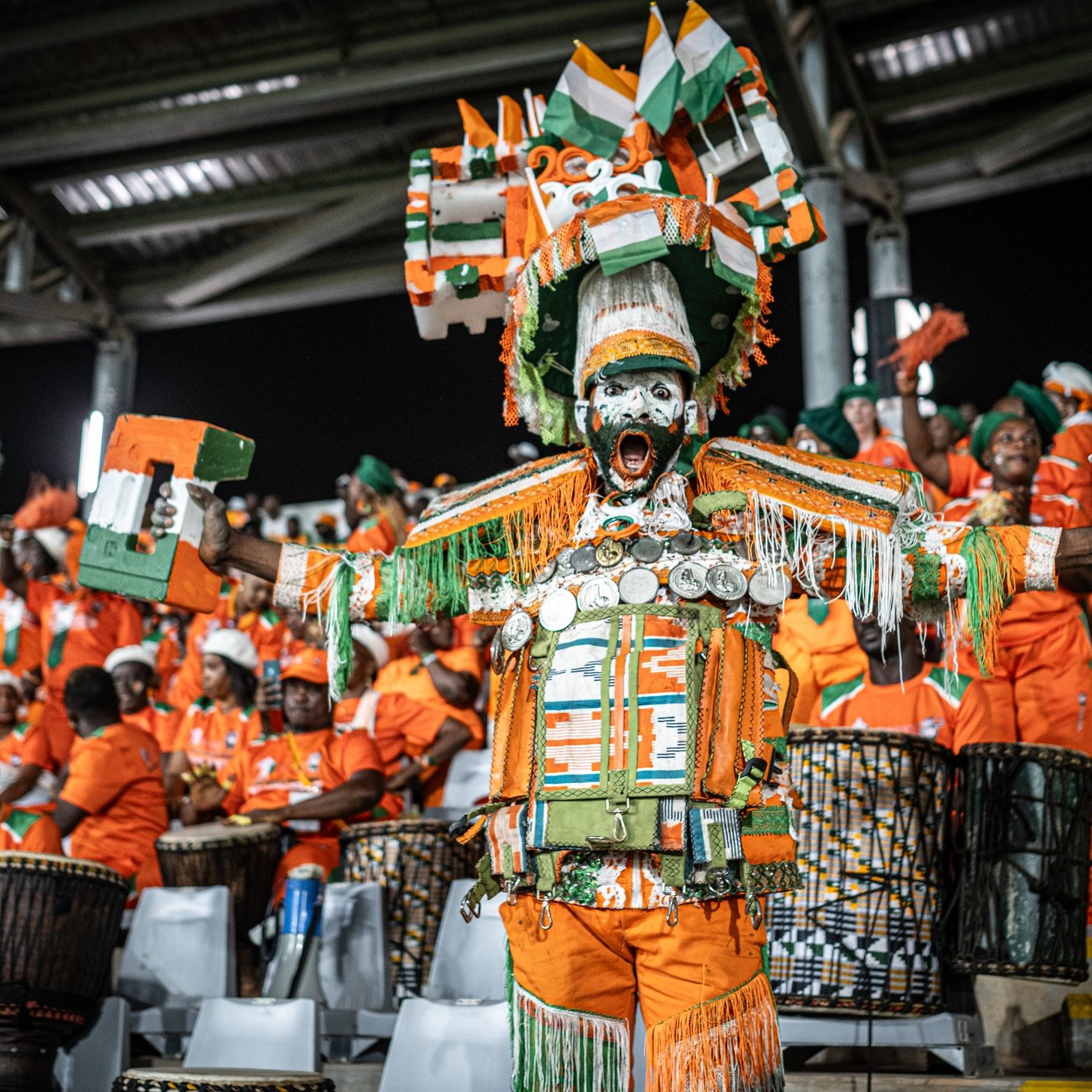 L'image montre une scène vibrante d'un événement festif, probablement un match de football. Au premier plan, un homme se distingue par son costume coloré composé de tissus orange et de nombreux accessoires décoratifs, dont des coquillages et des éléments en métal qui brillent à la lumière. Il est entouré de groupes de personnes, également en orange, qui jouent des tambours et sont vêtus de vêtements similaires. Tous semblent animés et enthousiastes, contribuant à une atmosphère de fête collective, ponctuée de chants et de danses. Les visages sont exprimés par des sourires et de la joie, avec des éléments traditionnels mettant en avant la culture locale.