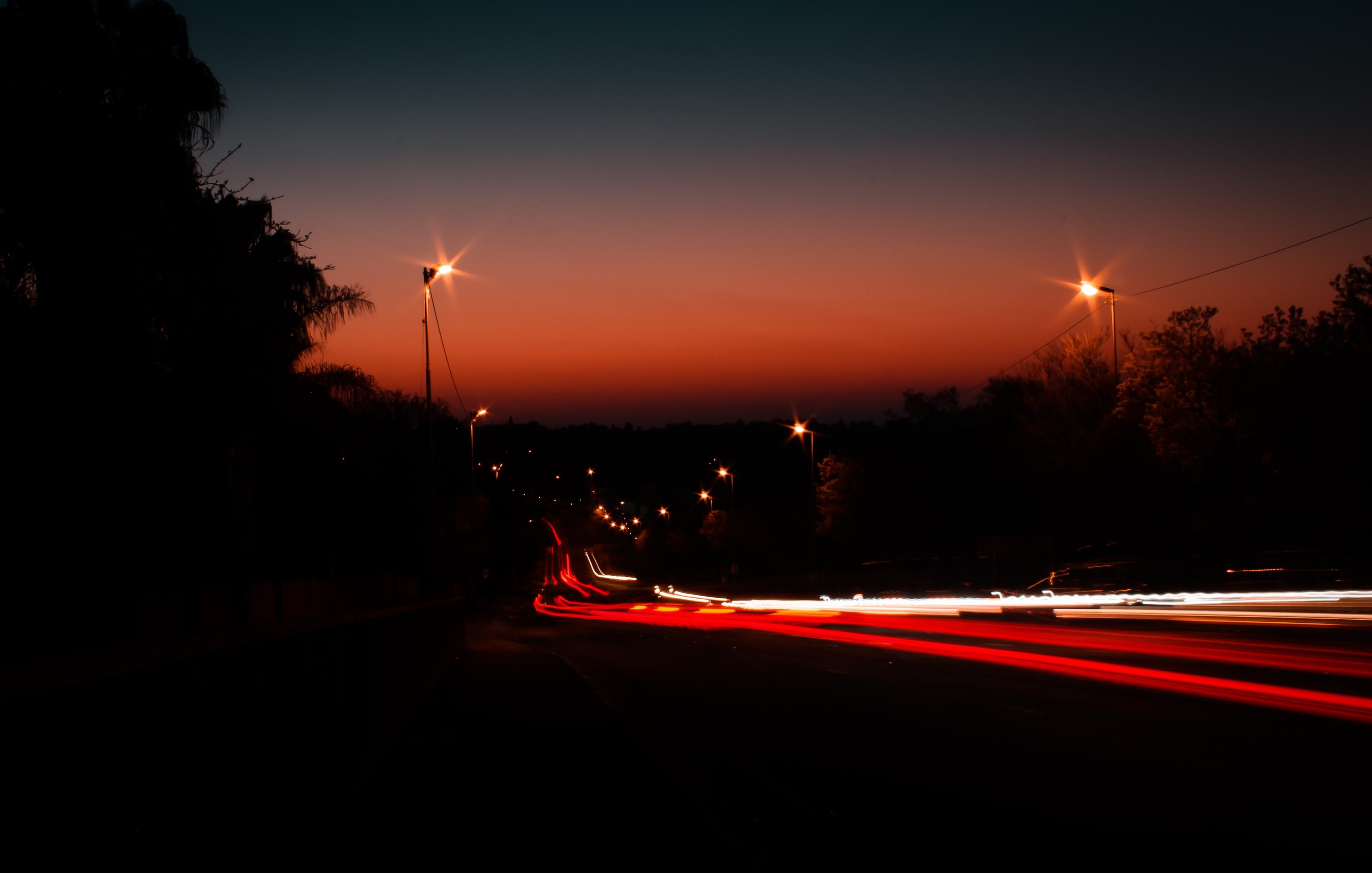 Imagine une scène tranquille à l'heure où le jour se transforme en nuit. Le ciel se teinte de dégradés d'orange et de bleu sombre, créant une ambiance douce et apaisante. Au loin, on aperçoit une route bordée d'arbres, presque silhouettes noires se découpant à l'horizon. Des lampadaires illuminent le chemin, leurs lumières jaunies ajoutant une touche chaleureuse à la scène. Des traces de lumière, comme des lignes fluides et vives, suggèrent des voitures qui passent rapidement, laissant derrière elles des filaments rouges et blancs. L'ensemble crée une atmosphère calme, invitant à la contemplation de ce moment transitionnel entre le jour et la nuit.