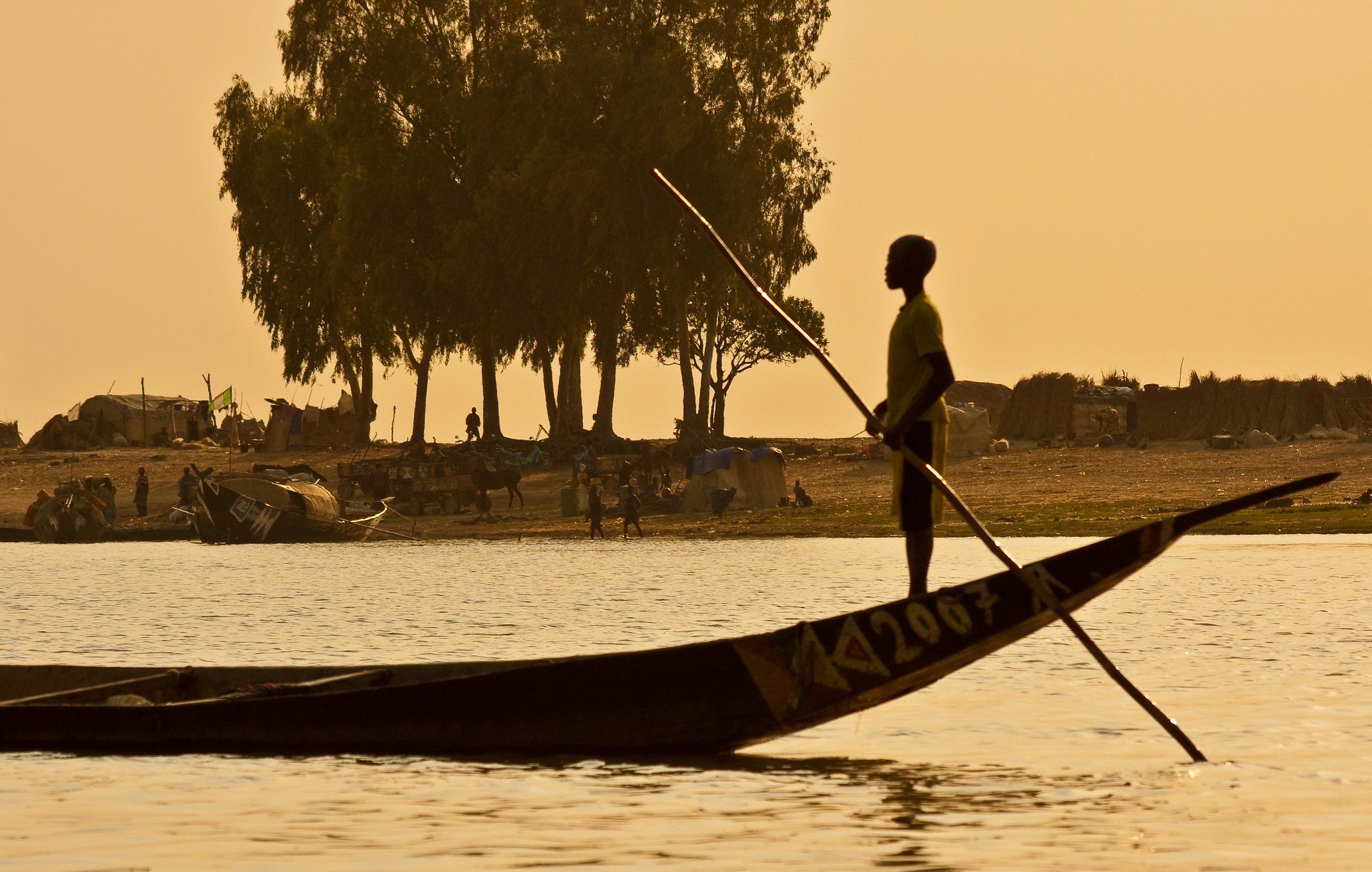 Imaginez une scène paisible au bord d'un fleuve. Un jeune garçon se tient debout dans une pirogue traditionnelle, les pieds ancrés sur le fond de la barque. Il utilise une longue perche pour diriger son embarcation à travers l'eau calme. En arrière-plan, on peut apercevoir un ruisseau de silhouettes d'arbres élancés, probablement des manguiers ou des sycomores, se découpant contre le ciel orangé du crépuscule. Sur la rive, des groupes de personnes s'affairent, tandis que des abris faits de toile et de paille sont visibles, suggérant une vie communautaire active. L'atmosphère est tranquille, teintée d'une chaleur douce, et l'ensemble dégage une sensation de sérénité et de connexion à la nature.