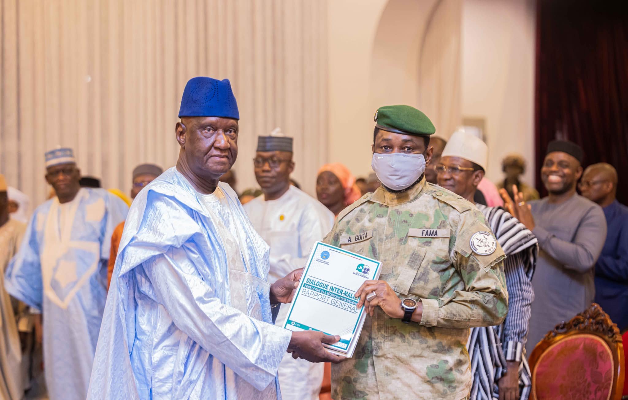 L'image montre un événement officiel ou une cérémonie de remise de prix. Au premier plan, un homme portant une tenue traditionnelle, composée d'une longue robe et d'un chapeau bleu, reçoit un document d'un officier militaire. Ce dernier est vêtu d'un uniforme camouflé et porte un masque gris. En arrière-plan, plusieurs personnes en tenue variée assistent à l'événement, certaines applaudissant, tandis que d'autres observent. L'environnement semble formel, avec des décorations adaptées à l'occasion.