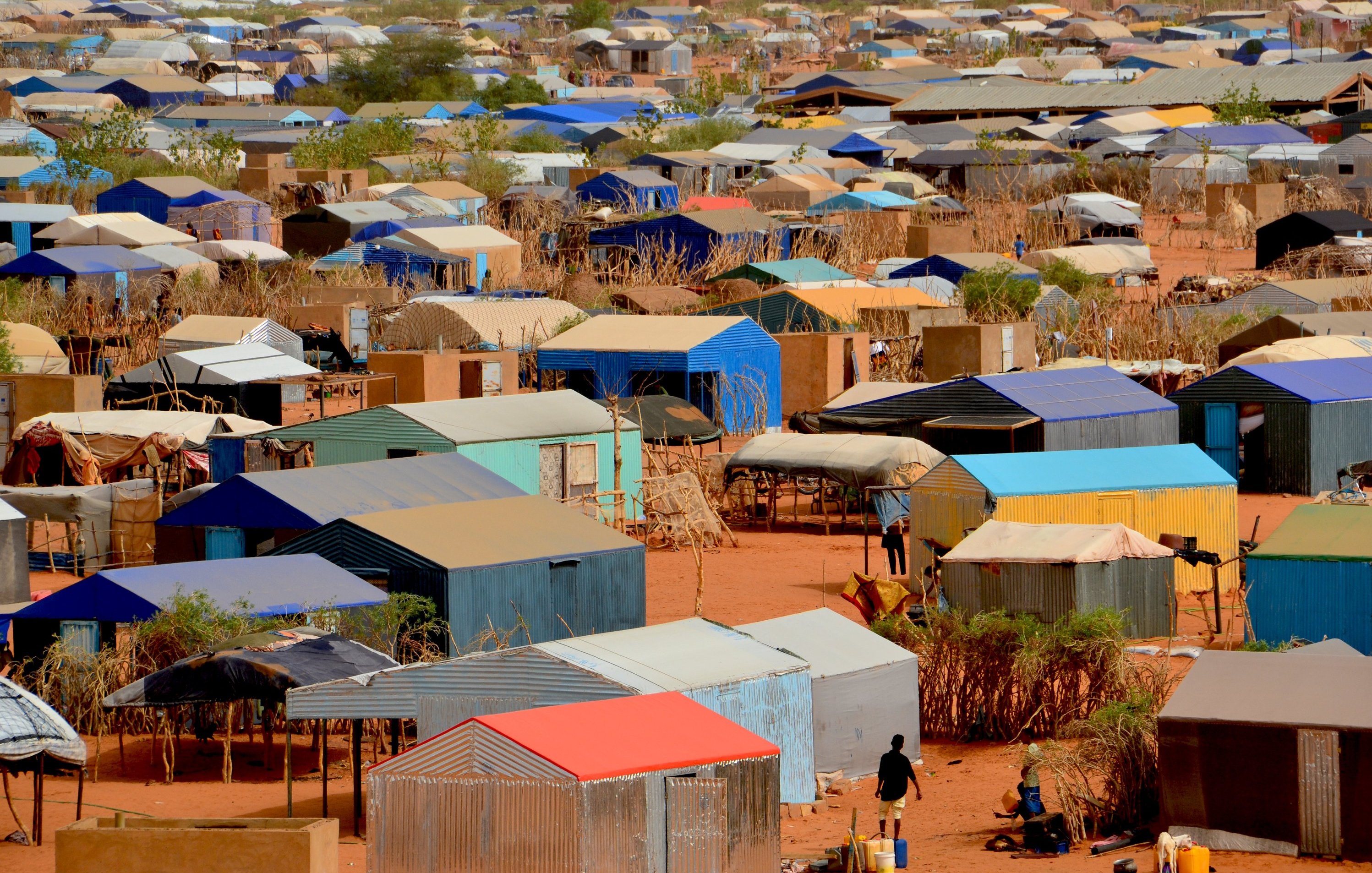 L'image montre un vaste camp de tentes et de logements temporaires disposés de manière dense sur un terrain désertique. Les structures sont recouvertes de toits de différentes couleurs, principalement des teintes de bleu, de rouge et de gris. Le sol est sablonneux, et quelques arbres ou buissons apparaissent ici et là, apportant un peu de verdure. En bas de l'image, on peut apercevoir des personnes se déplaçant entre les tentes, évoquant une activité quotidienne dans ce milieu. L'ensemble dégage une impression d'organisation dans la diversité des abris, tandis que le paysage aride souligne les conditions de vie dans ce camp.