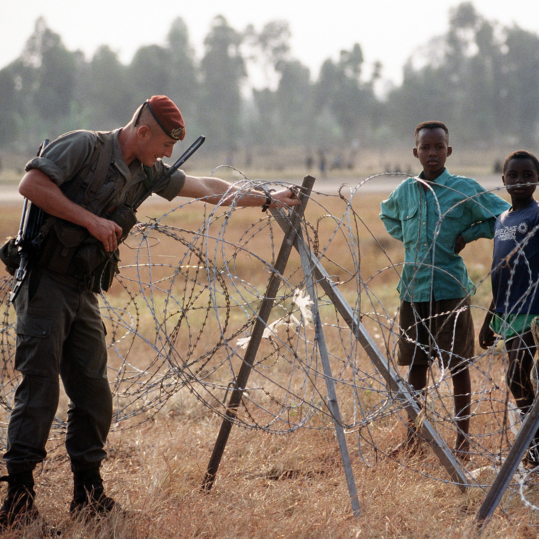 L'image présente un soldat en uniforme, concentré sur un fil barbelé, qui semble inspecter la barrière. Il est à proximité d'un groupe d'enfants qui l'observent. Les enfants, de différentes couleurs de vêtements, affichent des expressions variées, allant de la curiosité à l'inquiétude. L'arrière-plan montre un paysage ouvert, avec des essences d'arbres floues et des herbes sèches au sol. L'atmosphère dégage un mélange d'autorité et d'innocence, avec le soldat s'occupant de la sécurité, tandis que les enfants représentent l'innocence de l'enfance dans un contexte potentiellement difficile.