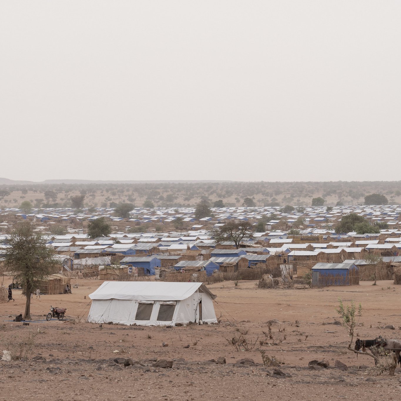 L'image montre un camp de réfugiés. Au premier plan, un enfant assis sur un amas de coussins et de matelas, portant un t-shirt rouge. Il semble pensif, regardant au loin. À côté de lui, un autre enfant est assis sur une petite table, vêtu d'un t-shirt bleu. L'environnement est poussiéreux, avec plusieurs tentes blanches en arrière-plan. On peut également apercevoir des objets éparpillés, comme des cartons et des bidons, évoquant une vie temporaire et difficile. L'atmosphère semble mélancolique et montre une réalité de vulnérabilité.