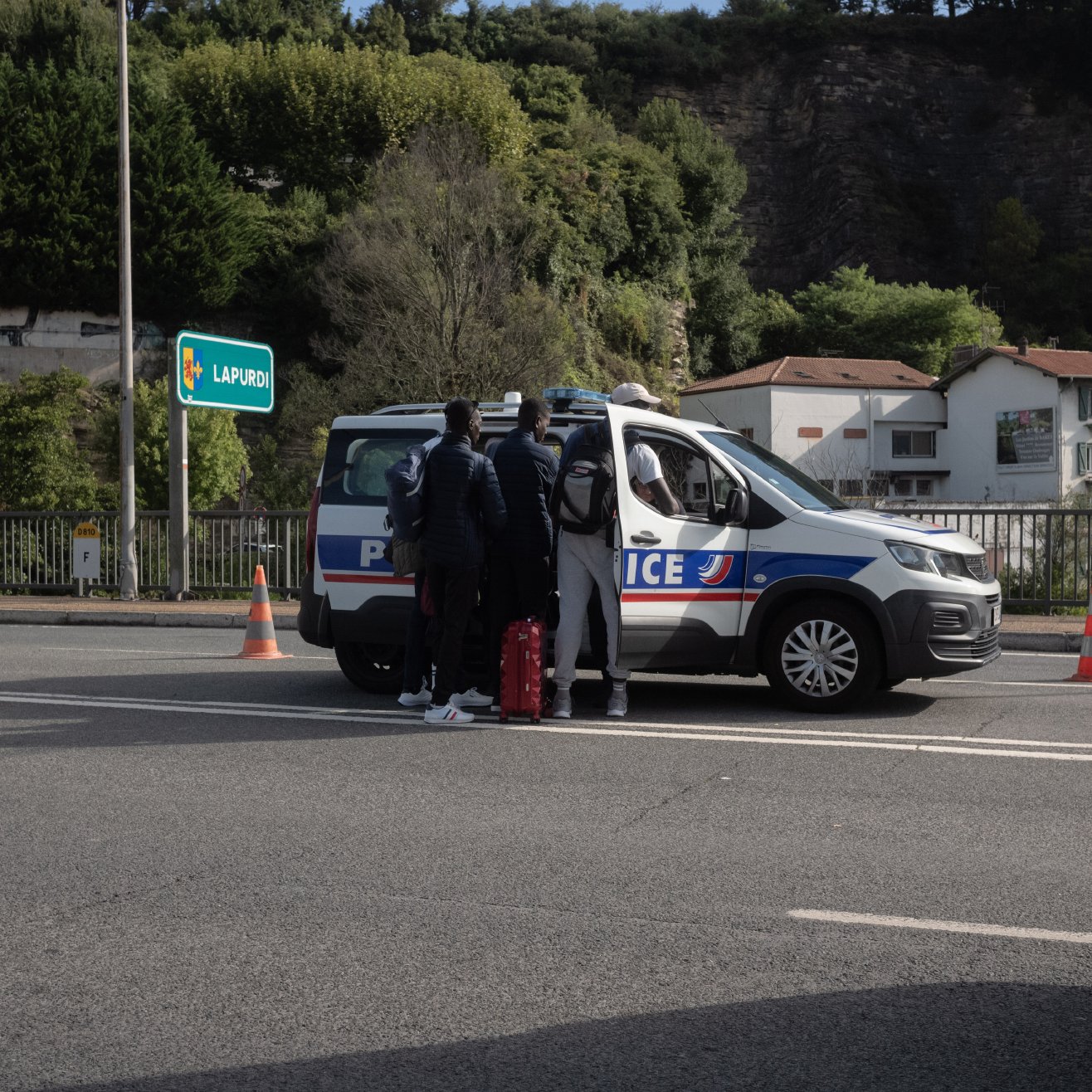 L'image montre une scène sur une route, probablement un contrôle de police. Au centre, on voit une voiture de police de couleur grise avec des marquages bleus et rouges, arrêtée sur la route. Quatre personnes se tiennent près de la voiture, dont deux semblent discuter avec les agents. Ils portent des vêtements sombres et certains semblent avoir des bagages. En arrière-plan, on aperçoit des bâtiments blancs et des arbres qui bordent la route, avec un panneau indiquant "LARRESO". Des cônes de signalisation orange sont disposés autour de la voiture pour sécuriser la zone. L'ambiance paraît calme, bien que la situation soit formelle.