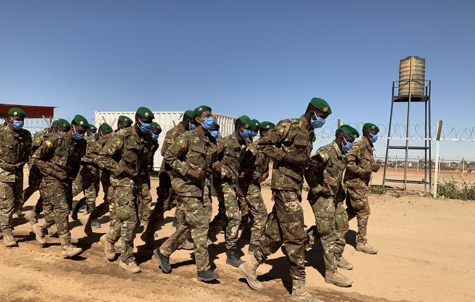 Dans cette image, un groupe de soldats est en train de marcher en formation sur un chemin poussiéreux. Ils portent des uniformes militaires verts camouflés et des casquettes vertes. La majorité d'entre eux portent également des masques bleus, ce qui suggère une mesure de protection sanitaire. En arrière-plan, on peut distinguer des conteneurs et une tour d'eau. Le ciel est dégagé, avec une couleur bleue intense, créant un contraste avec la terre orange et les silhouettes des soldats. L'atmosphère évoque un moment de discipline et d'organisation dans un environnement militaire.