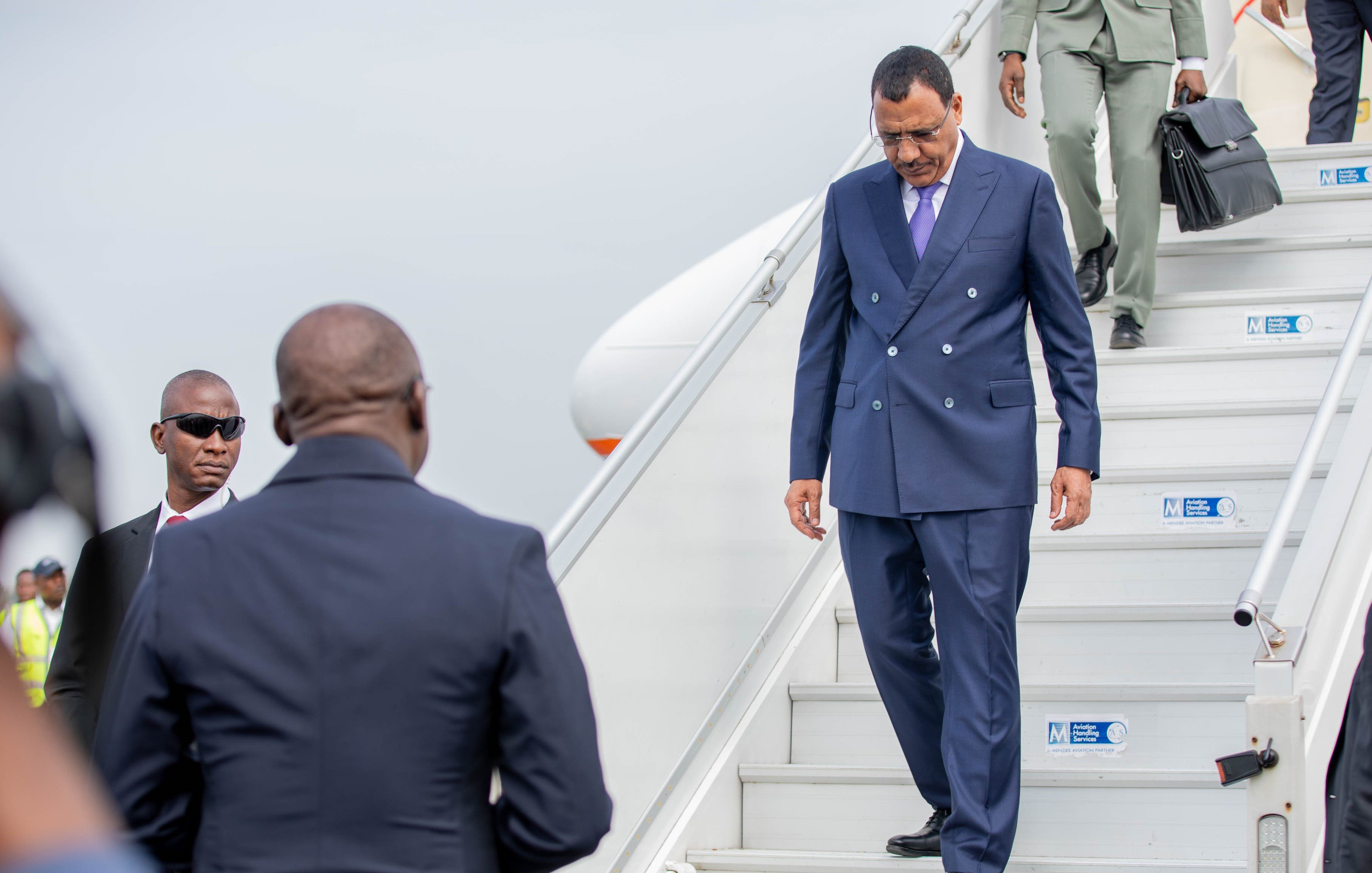 L'image montre un homme qui descend des marches d'un avion. Il porte un costume bleu foncé et des lunettes de soleil. Son expression semble concentrée tandis qu'il s'avance avec assurance. À côté de lui, un autre homme en costume noir observe attentivement. En arrière-plan, on aperçoit des membres du personnel de l'aéroport, ainsi que l'avion stationné. L'atmosphère peut sembler formelle et dynamique, suggérant un moment important, vraisemblablement une arrivée ou une sortie officielle.