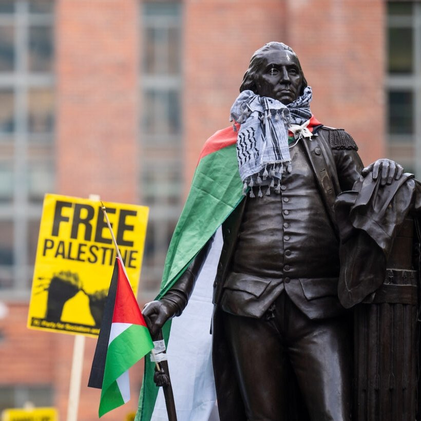 L'image montre une statue en bronze d'un homme debout, tenant un bâton dans une main et un document dans l'autre. Cette statue est habillée d'un keffieh, un foulard traditionnel palestinien en noir et blanc, et elle est drapée d'un drapeau palestinien au motif rouge, blanc, noir et vert. En arrière-plan, on aperçoit un panneau jaune sur lequel est inscrit "FREE PALESTINE" en lettres noires, exprimant un message de soutien pour la cause palestinienne. L'environnement semble être une place publique animée, avec des bâtiments en briques visibles derrière. Cette scène témoigne de la mobilisation et de la solidarité, créant une ambiance engageante et militante.